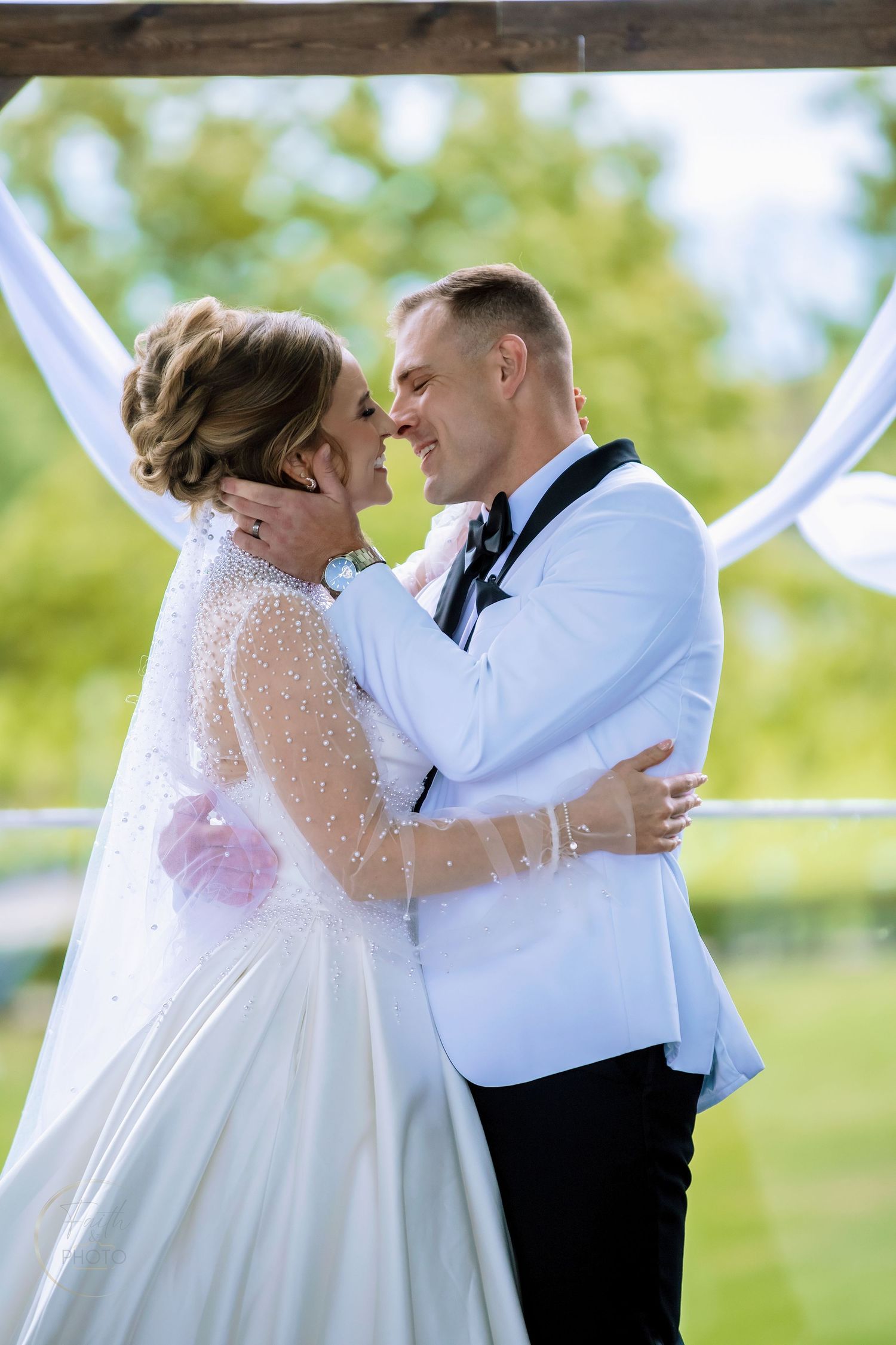 A bride and groom share a romantic embrace outdoors at their wedding ceremony, surrounded by white draping and lush greenery.