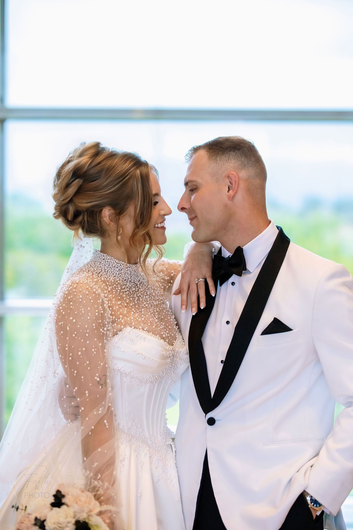 Bride and groom share a joyful moment, she in a pearl-adorned gown, he in a white tuxedo, gazing lovingly at each other.