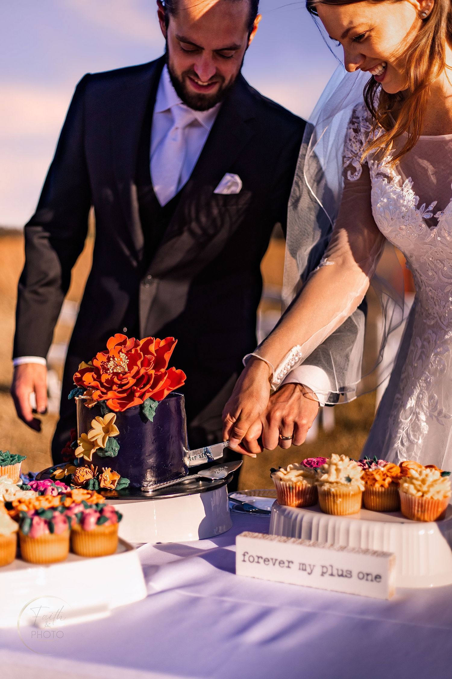 Bride and groom cutting a dark floral wedding cake surrounded by colorful cupcakes at an outdoor reception.