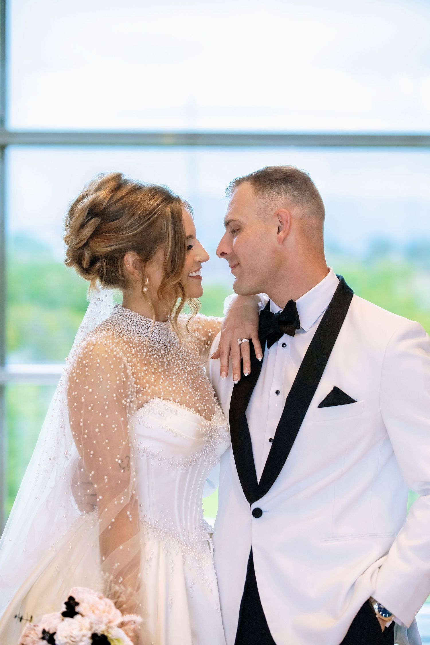 Bride and groom share a joyful moment, she in a pearl-adorned gown with veil, he in a white tuxedo, near large windows.