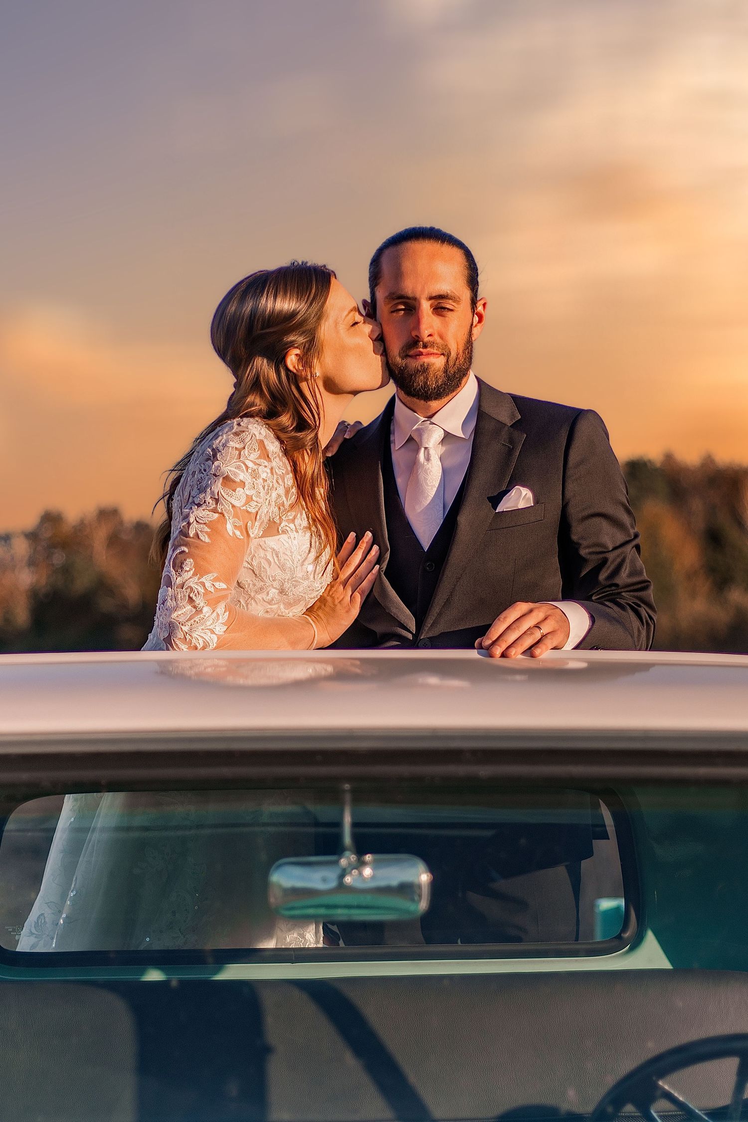 Bride kissing groom on cheek beside vintage white car at golden sunset, wedding couple in elegant attire outdoors.