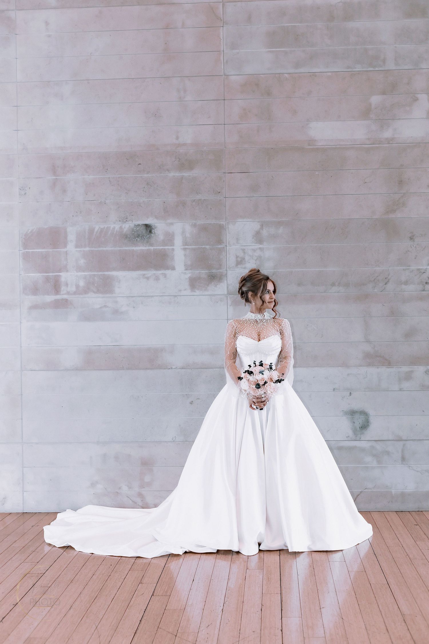 Elegant bride in a white ball gown with lace sleeves holding a bouquet, standing before a concrete wall.