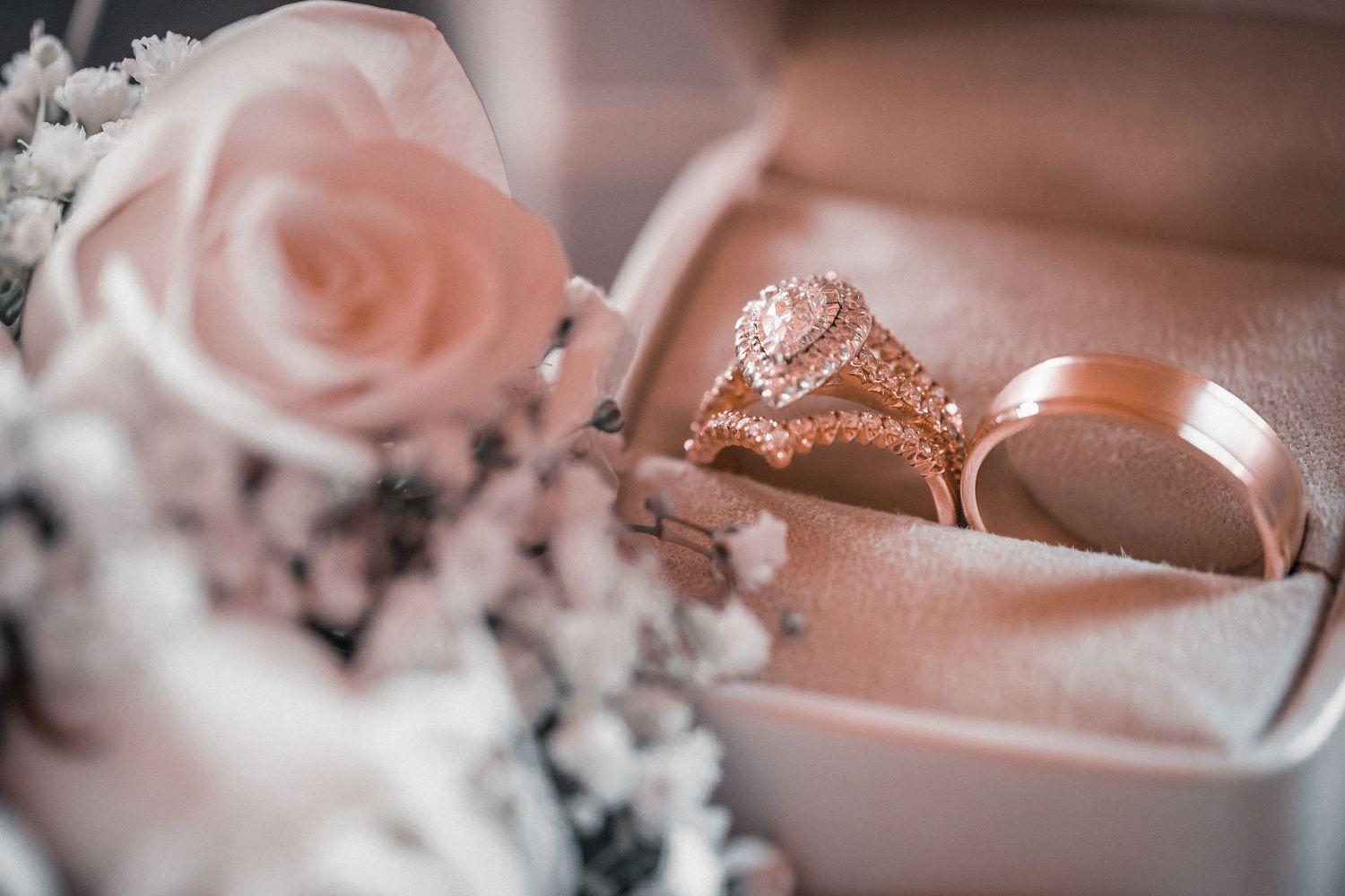 A rose gold engagement ring and wedding band nestled in a pink box with a blush rose and baby's breath flowers.