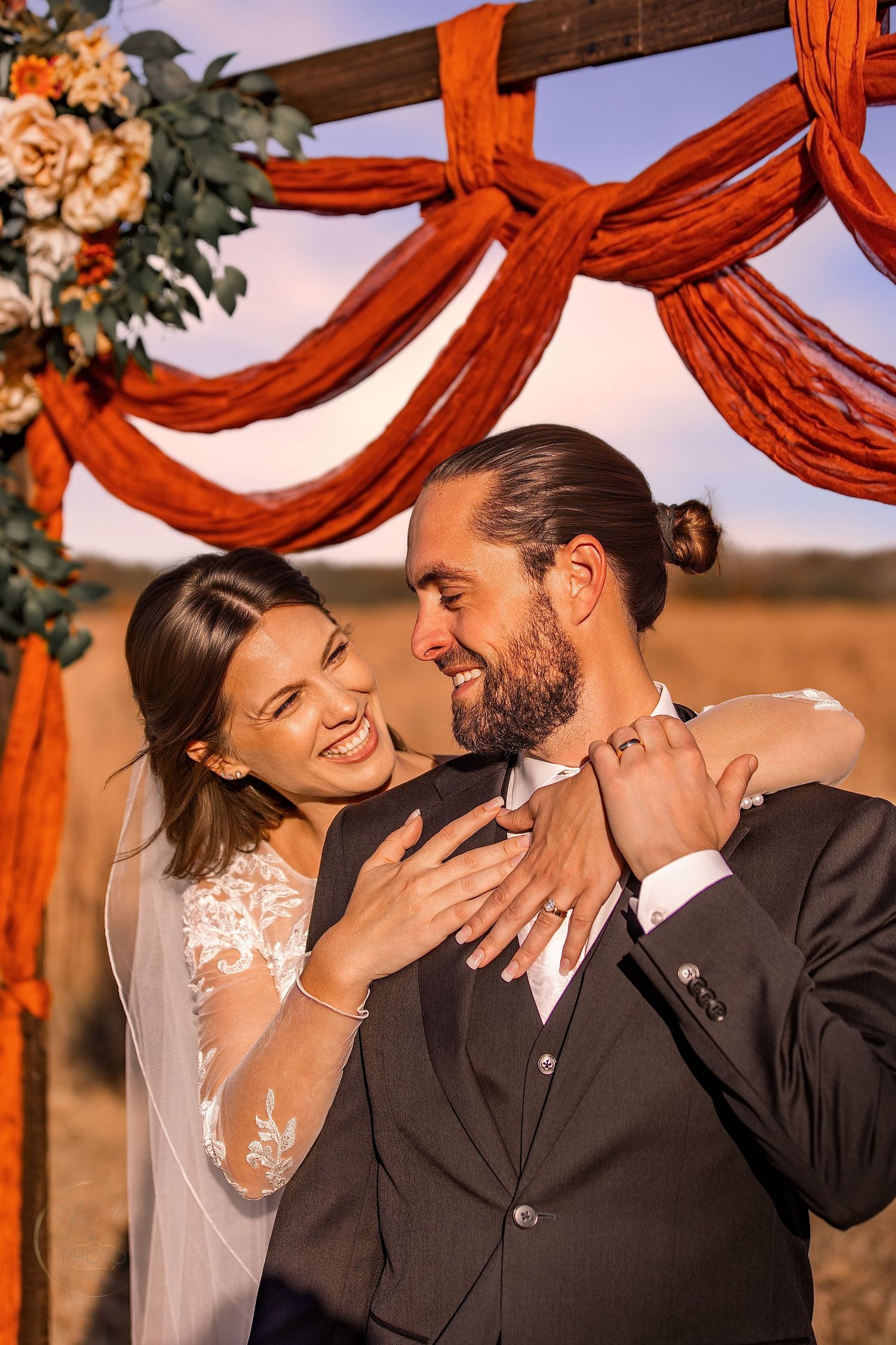 Happy newlyweds embracing under rustic orange draped wedding arch outdoors at golden hour.