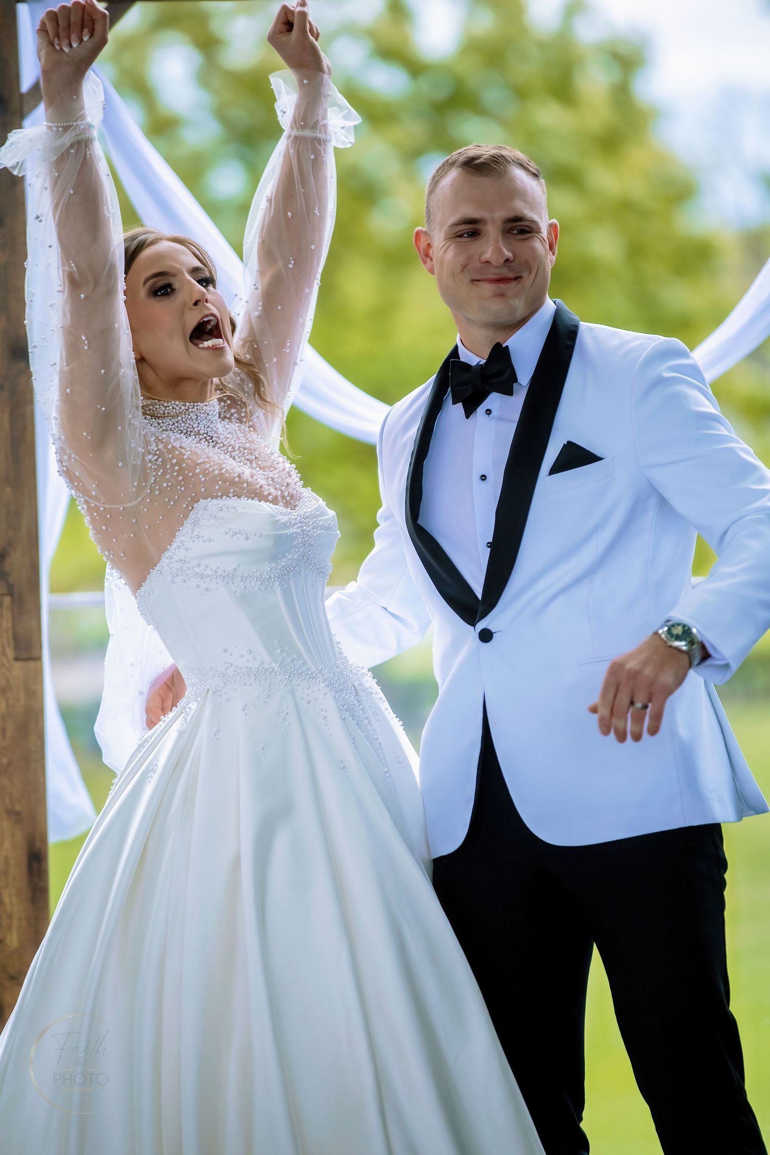 Joyful bride with arms raised and groom in white tuxedo celebrating at outdoor wedding ceremony.