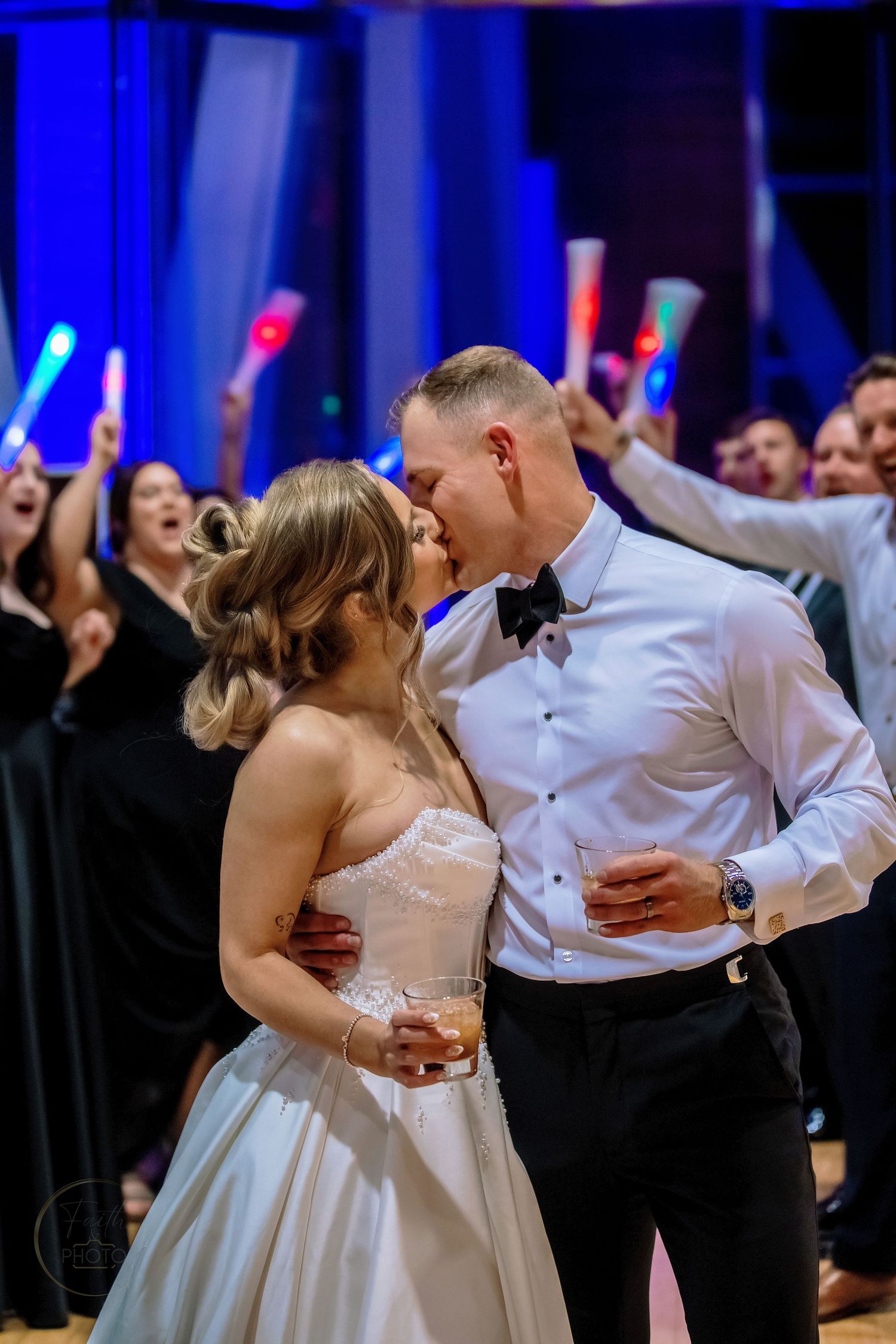 Bride and groom share a kiss on the dance floor surrounded by cheering guests holding colorful glow sticks at their wedding reception.