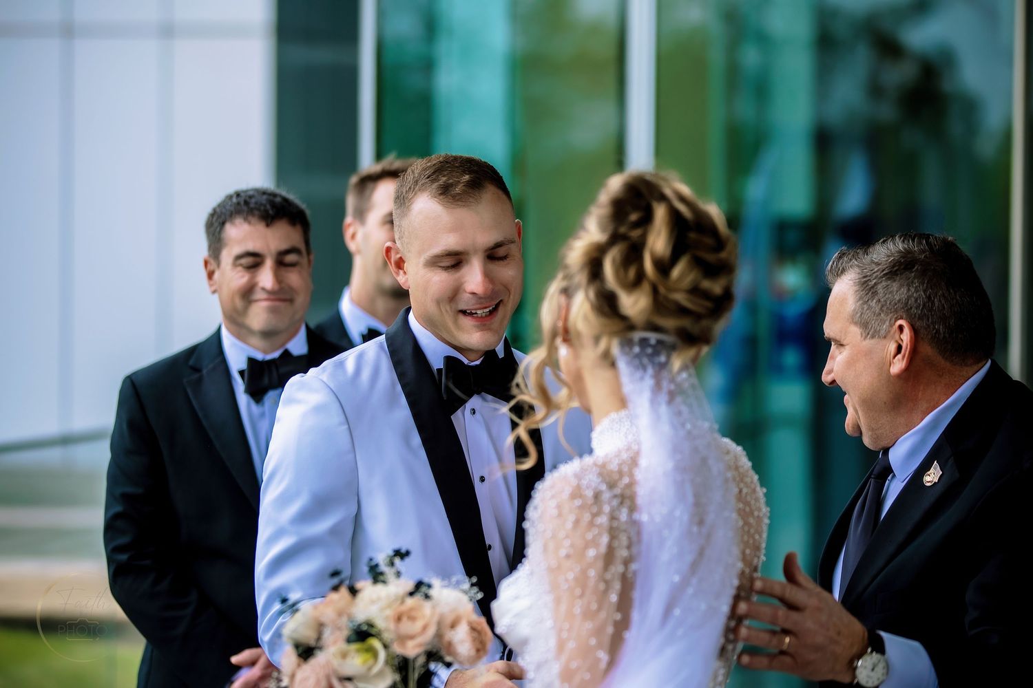 Groom in black bow tie smiles at bride holding bouquet, surrounded by guests outside modern building.