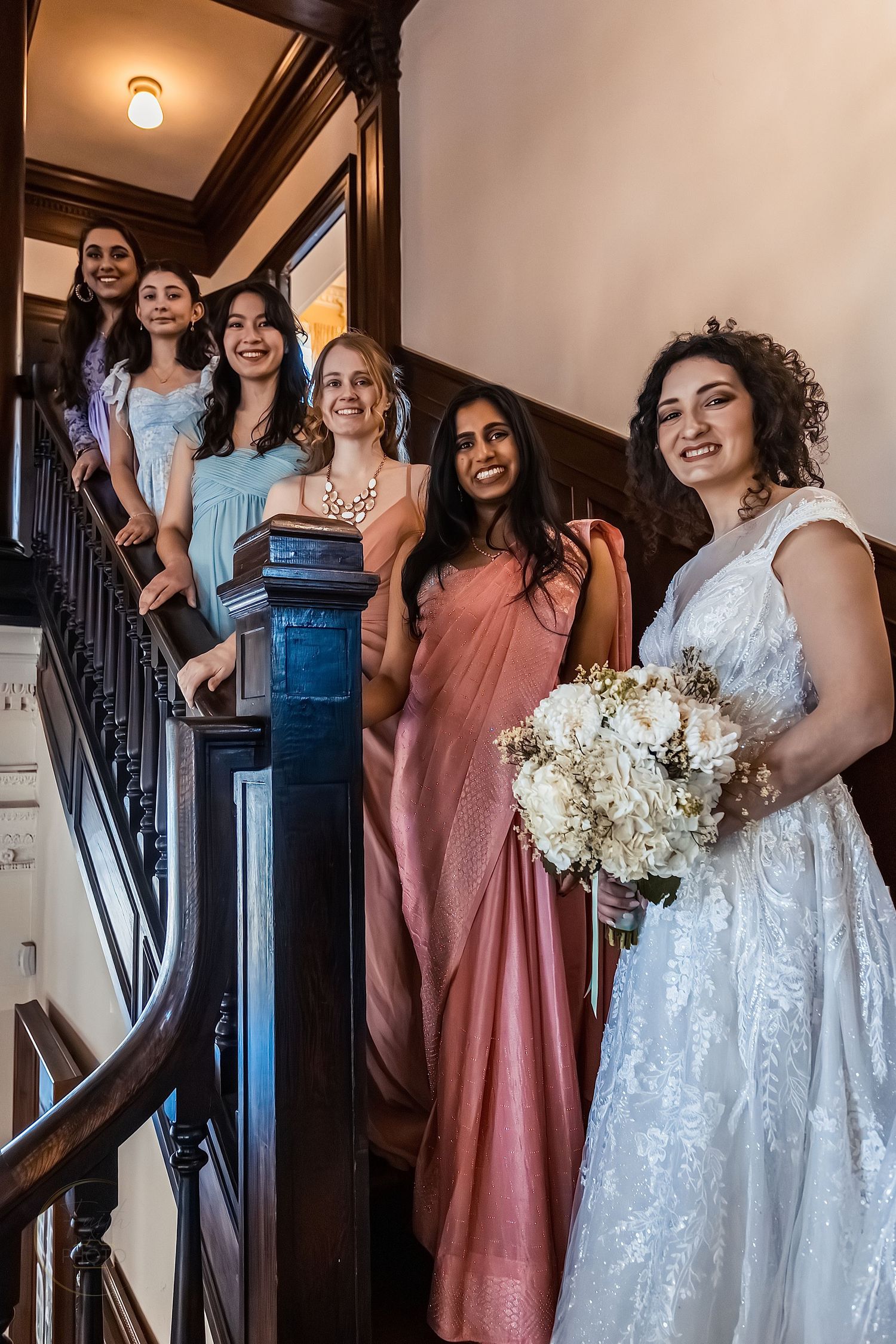 Bride in white gown holding bouquet poses with bridesmaids in colorful dresses on elegant wooden staircase.