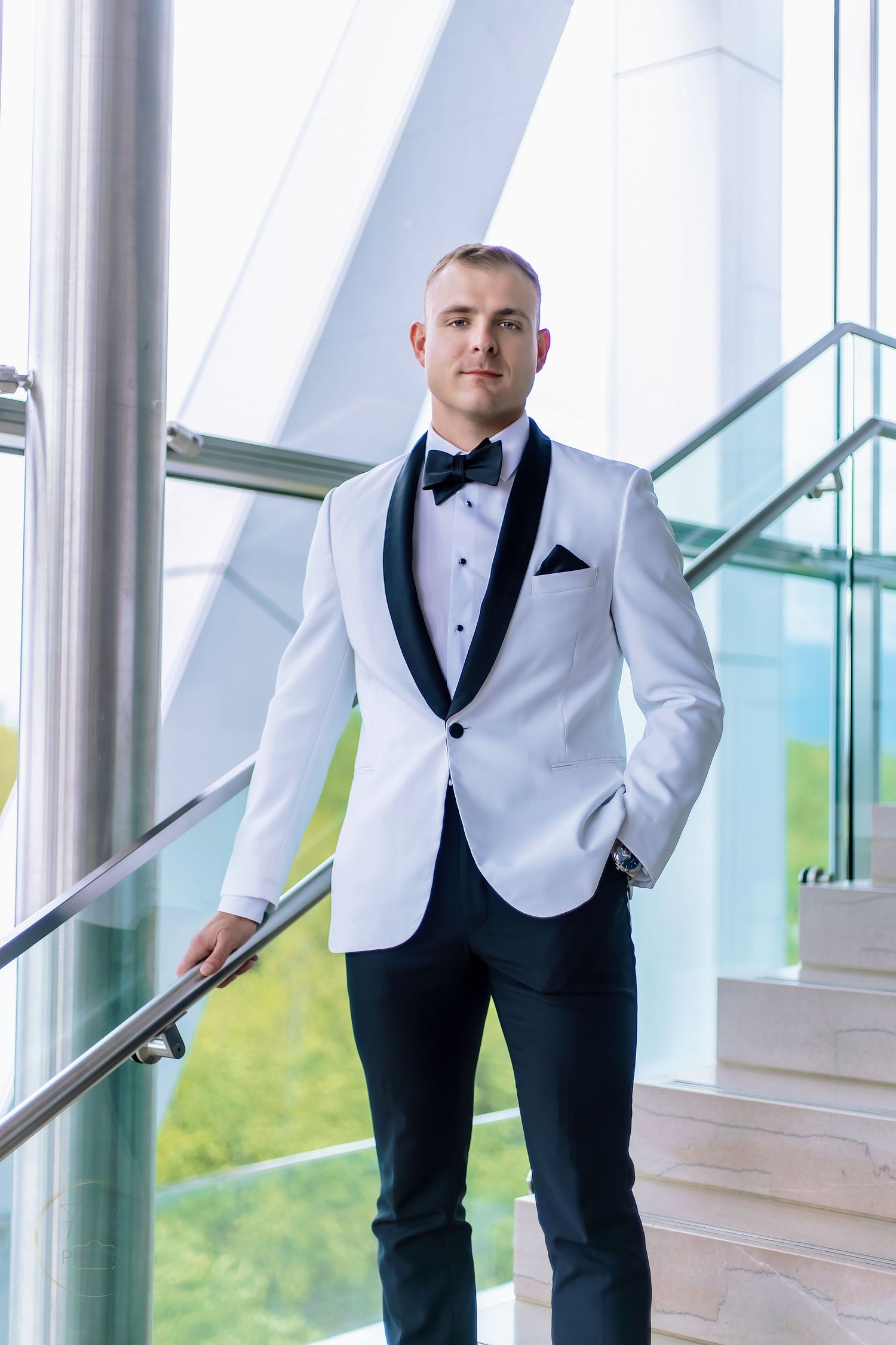 Man in white tuxedo jacket with black lapels and bow tie standing on stairs in modern building.