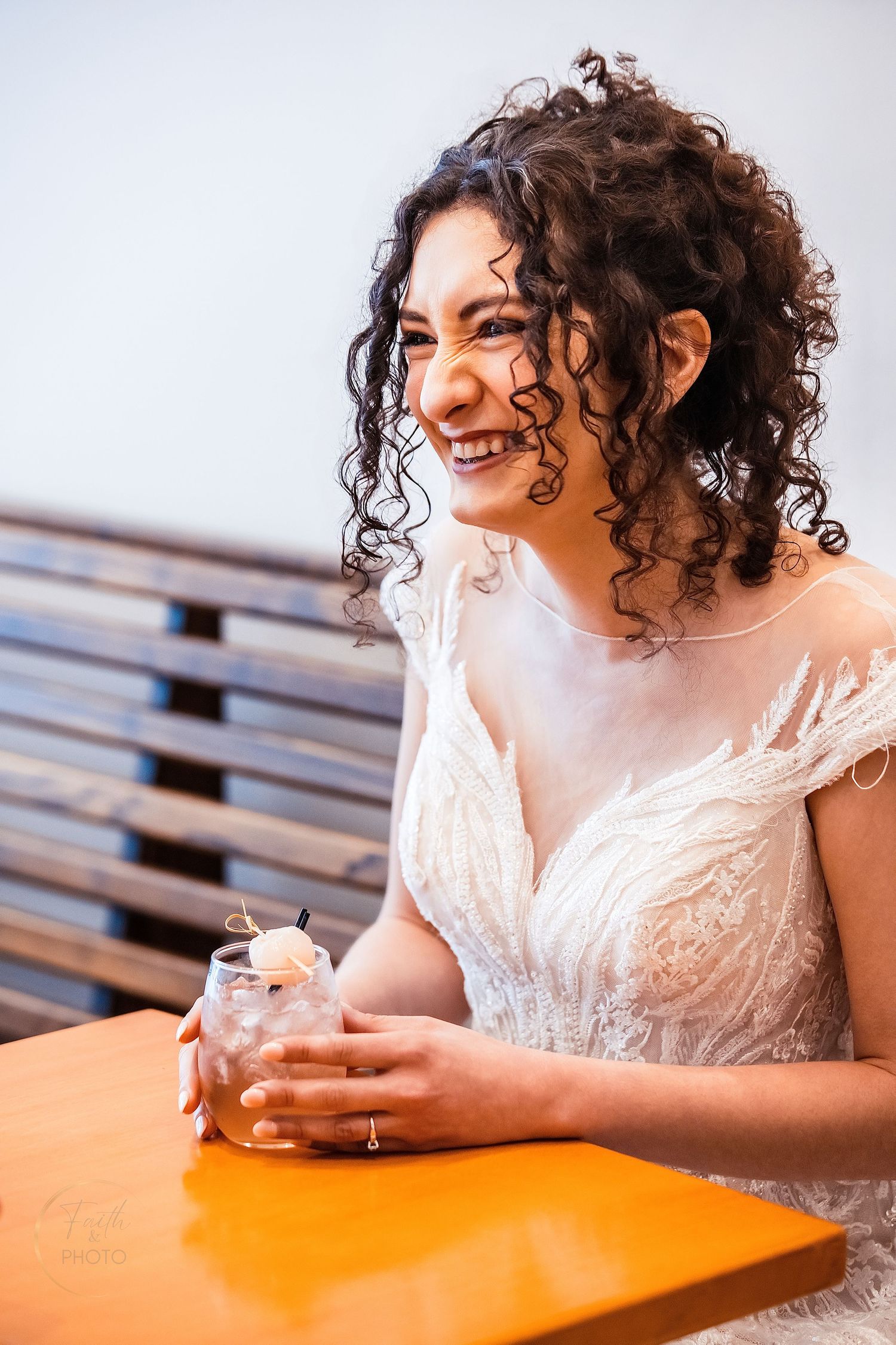 Smiling bride with curly hair in white lace wedding dress holding a drink at an orange table.