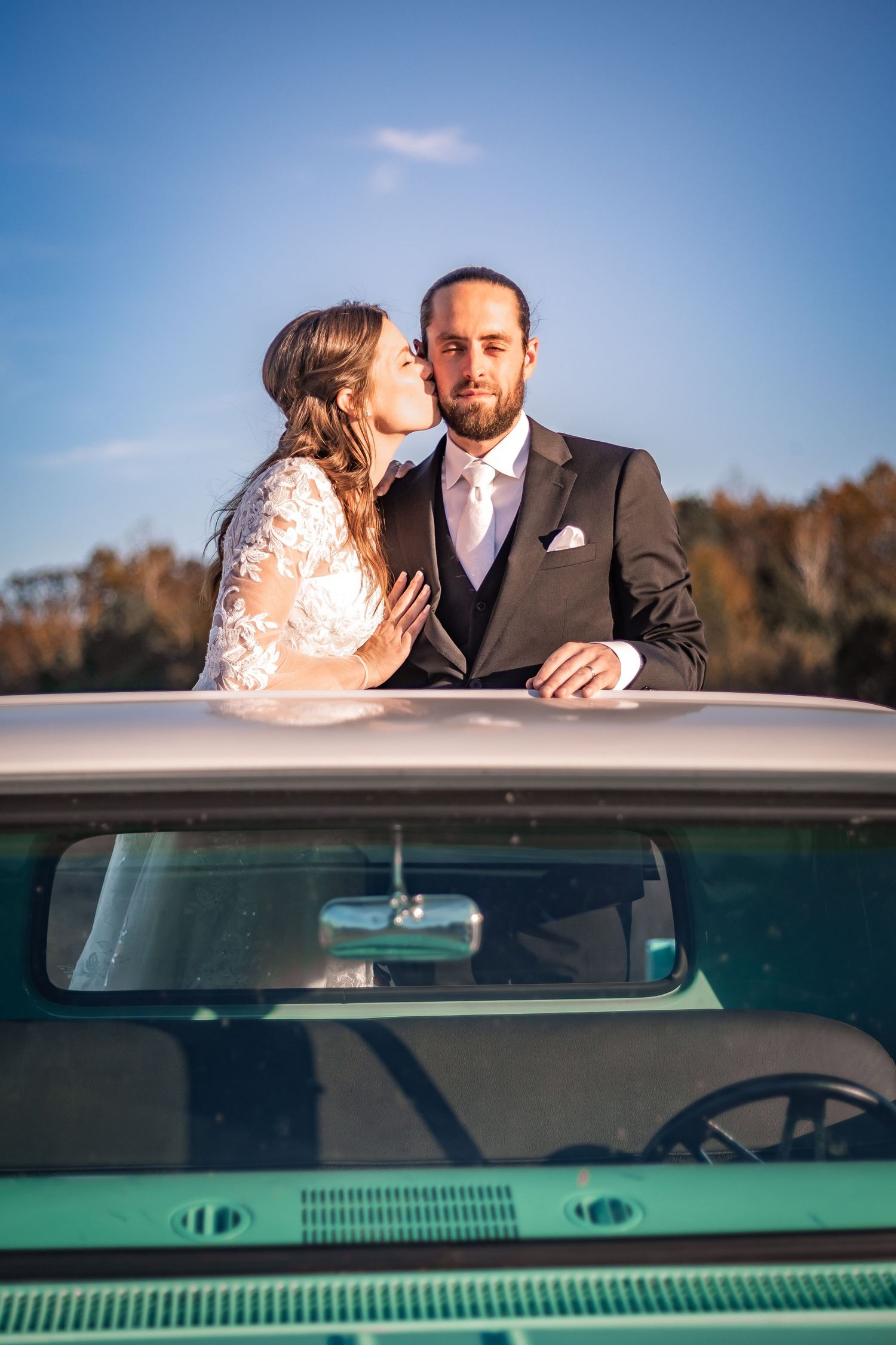 A romantic couple shares tender moments while seated in a vintage car on their wedding day against a blue sky background.