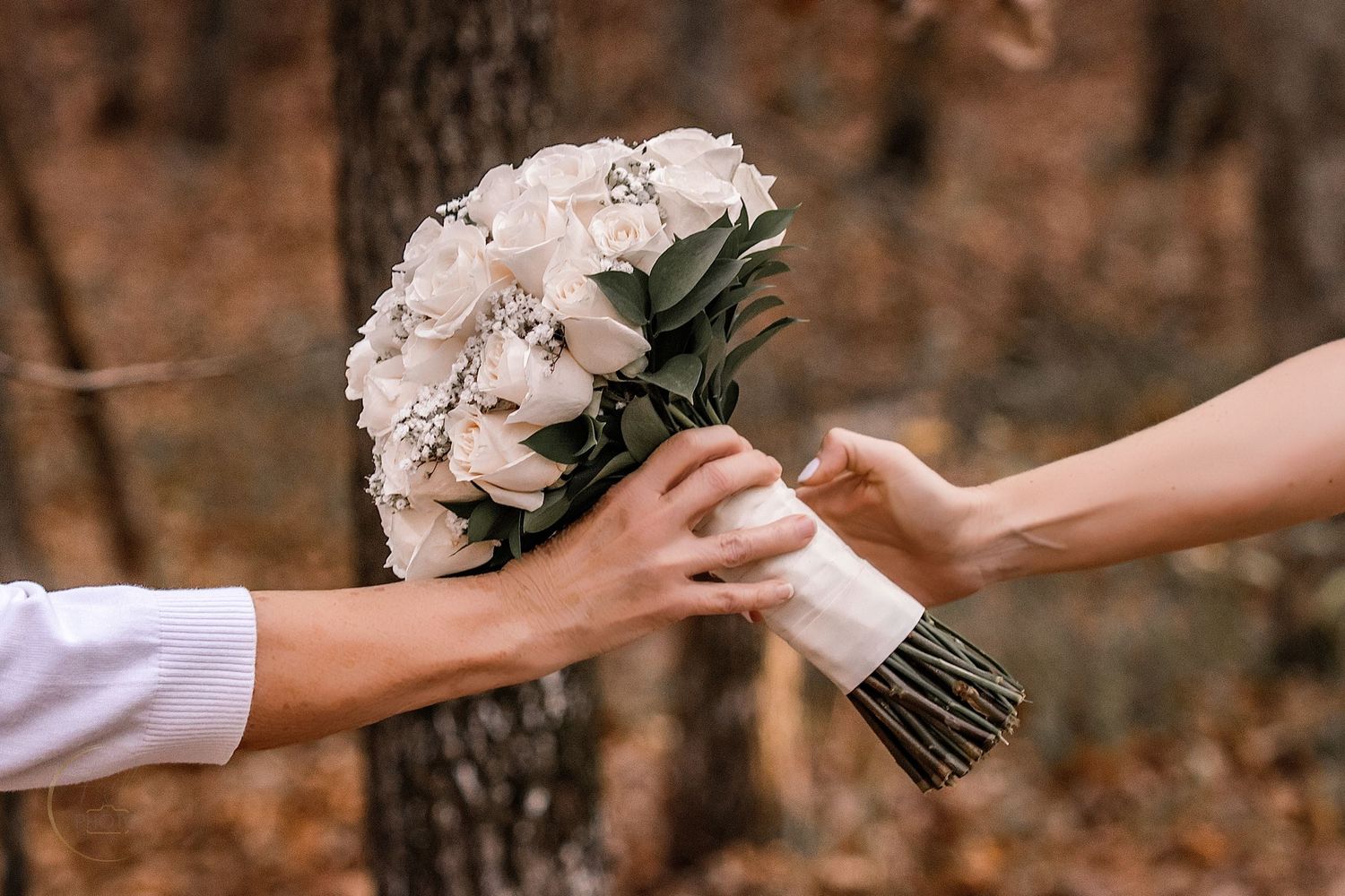 Two hands exchanging a white rose wedding bouquet in an autumn forest setting.