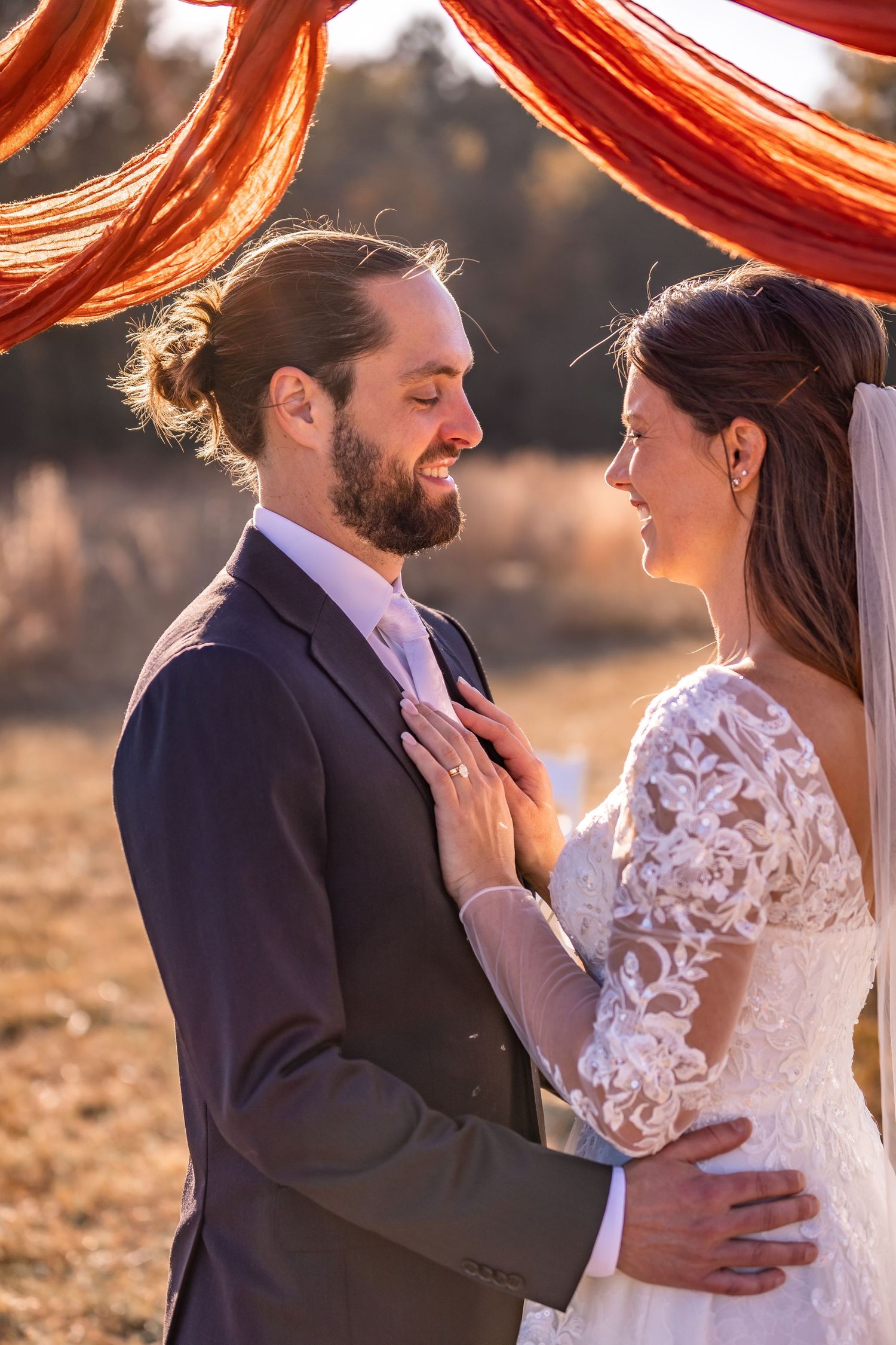 A romantic wedding couple embraces in golden sunlight wearing traditional formal attire with lace details.