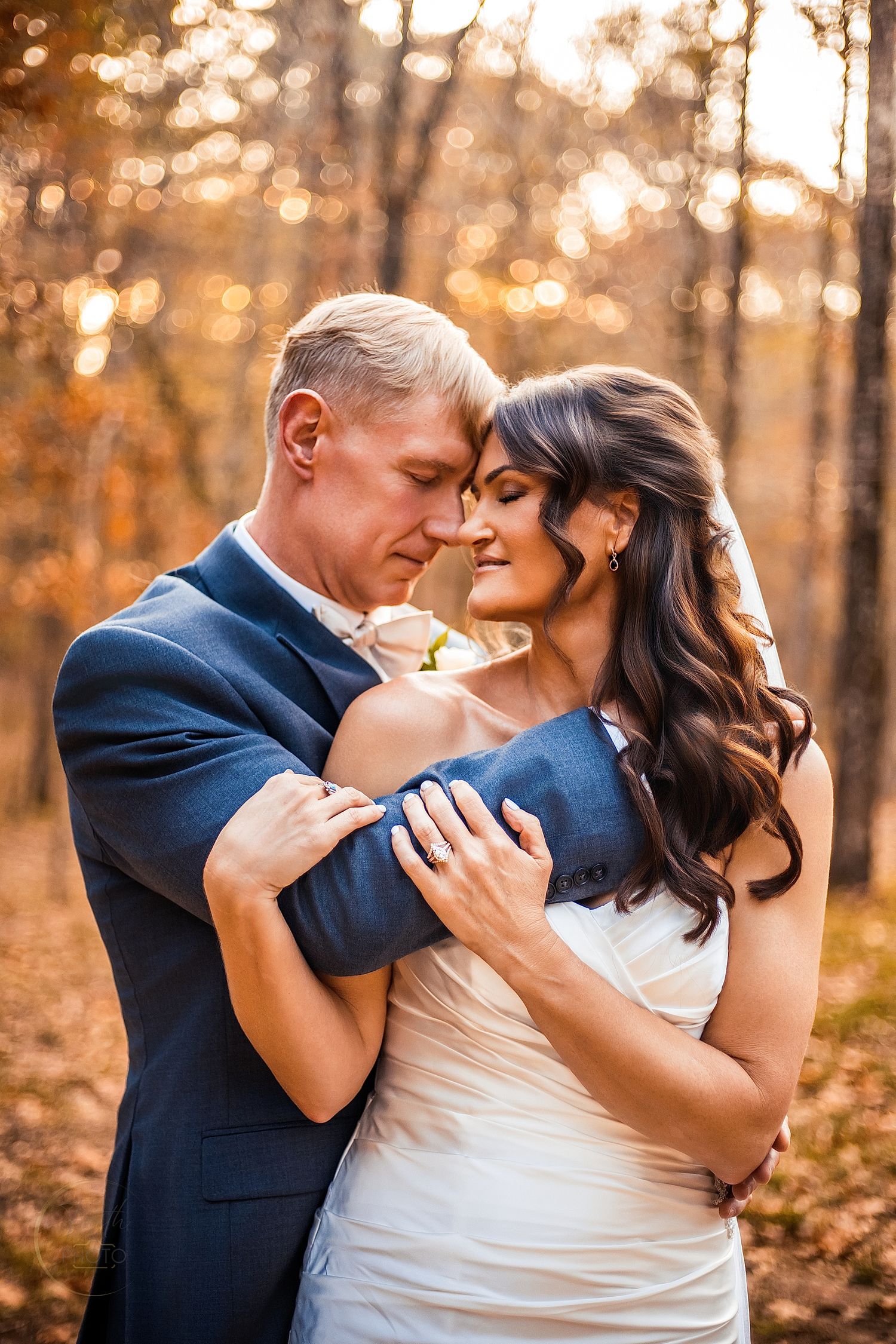Bride and groom embrace tenderly in an autumn forest, wearing white dress and navy suit, with golden bokeh light.