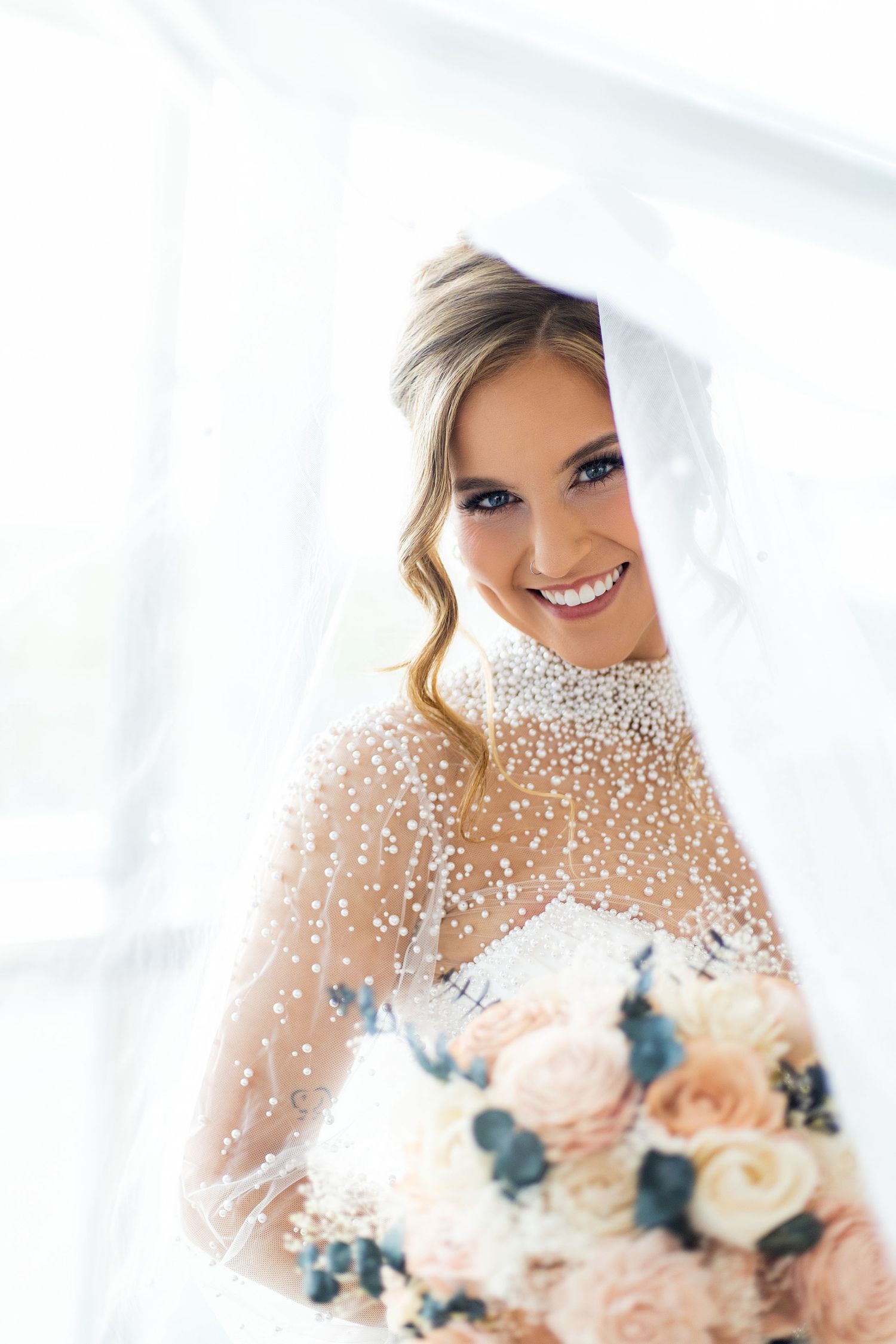 Smiling bride in pearl-adorned veil and gown holds a blush and white rose bouquet in a bright, airy setting.
