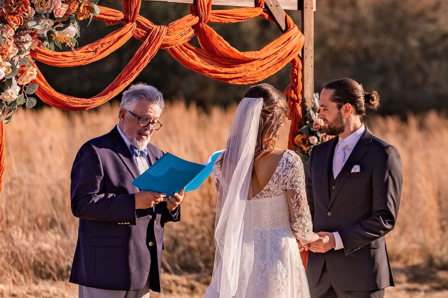 Outdoor wedding ceremony with officiant reading from blue folder, bride in lace gown, groom in suit, beneath orange floral arch.