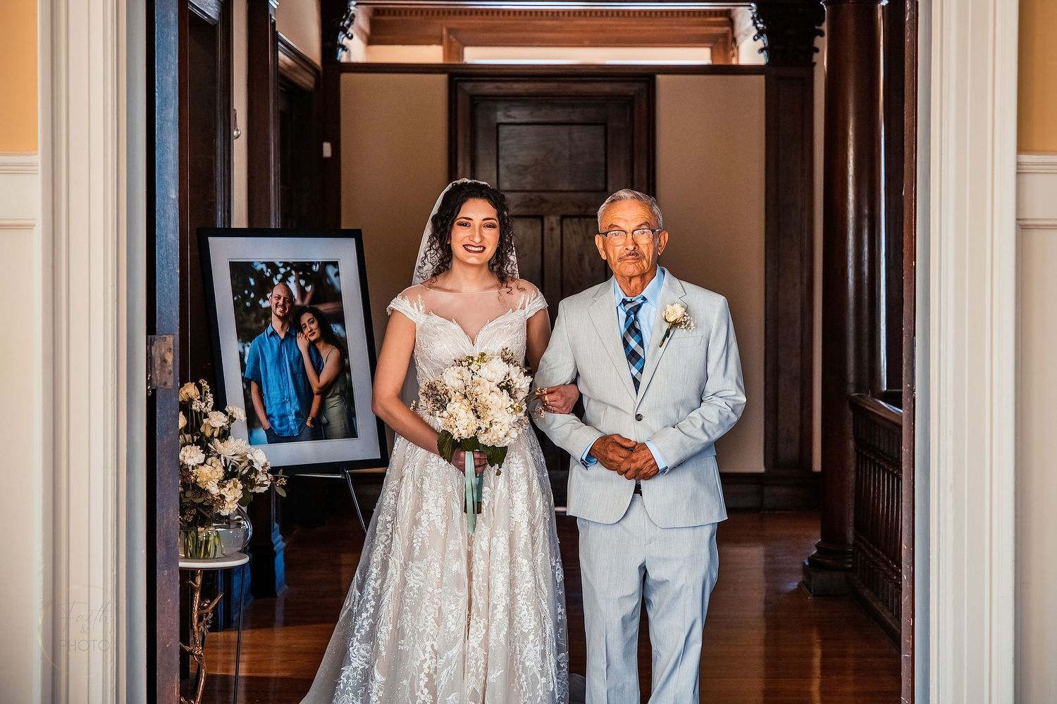 Bride in lace gown walks down aisle with elderly man in gray suit, bouquet in hand, framed photo beside them.