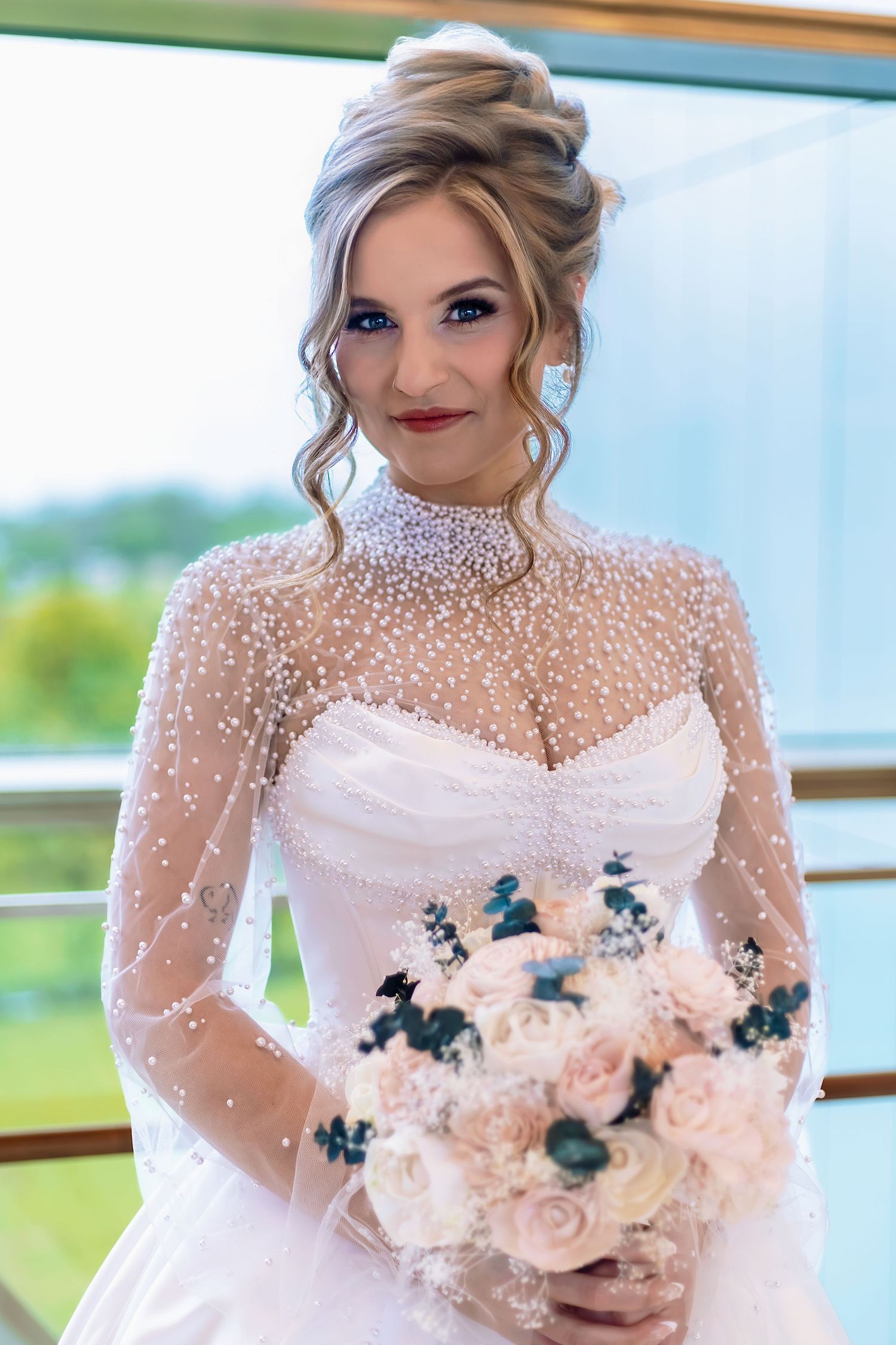 Elegant bride in pearl-embellished white gown holding blush rose bouquet, wearing updo hairstyle and dramatic makeup.