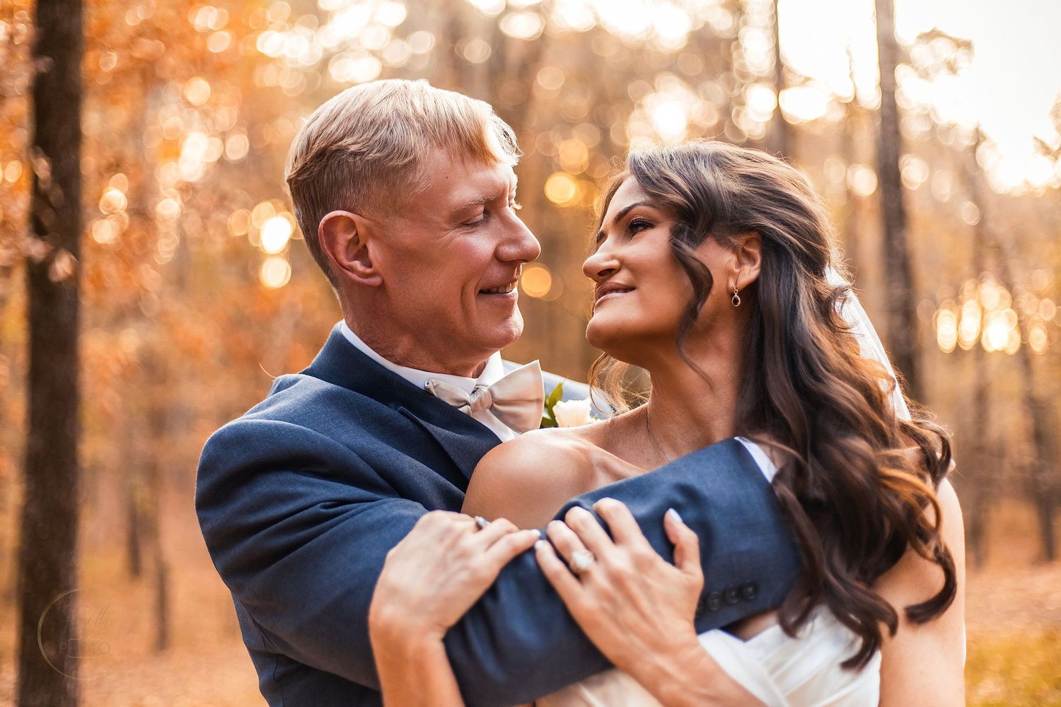 A couple embraces in an autumn forest, the groom in a navy suit and bride in white, sharing a romantic wedding moment.