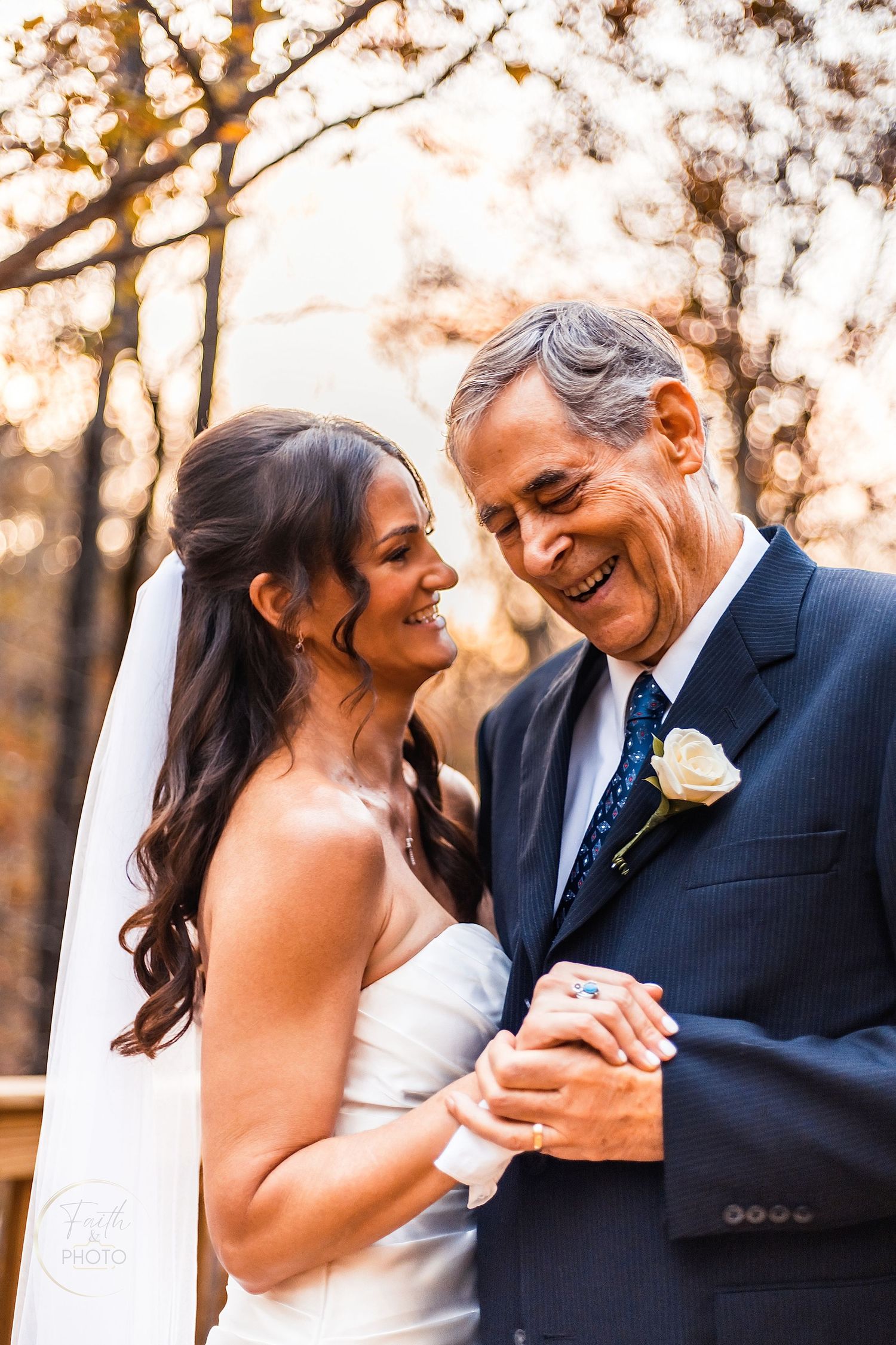 Bride in white gown and veil shares a joyful moment with older man in navy suit with boutonniere at outdoor wedding.
