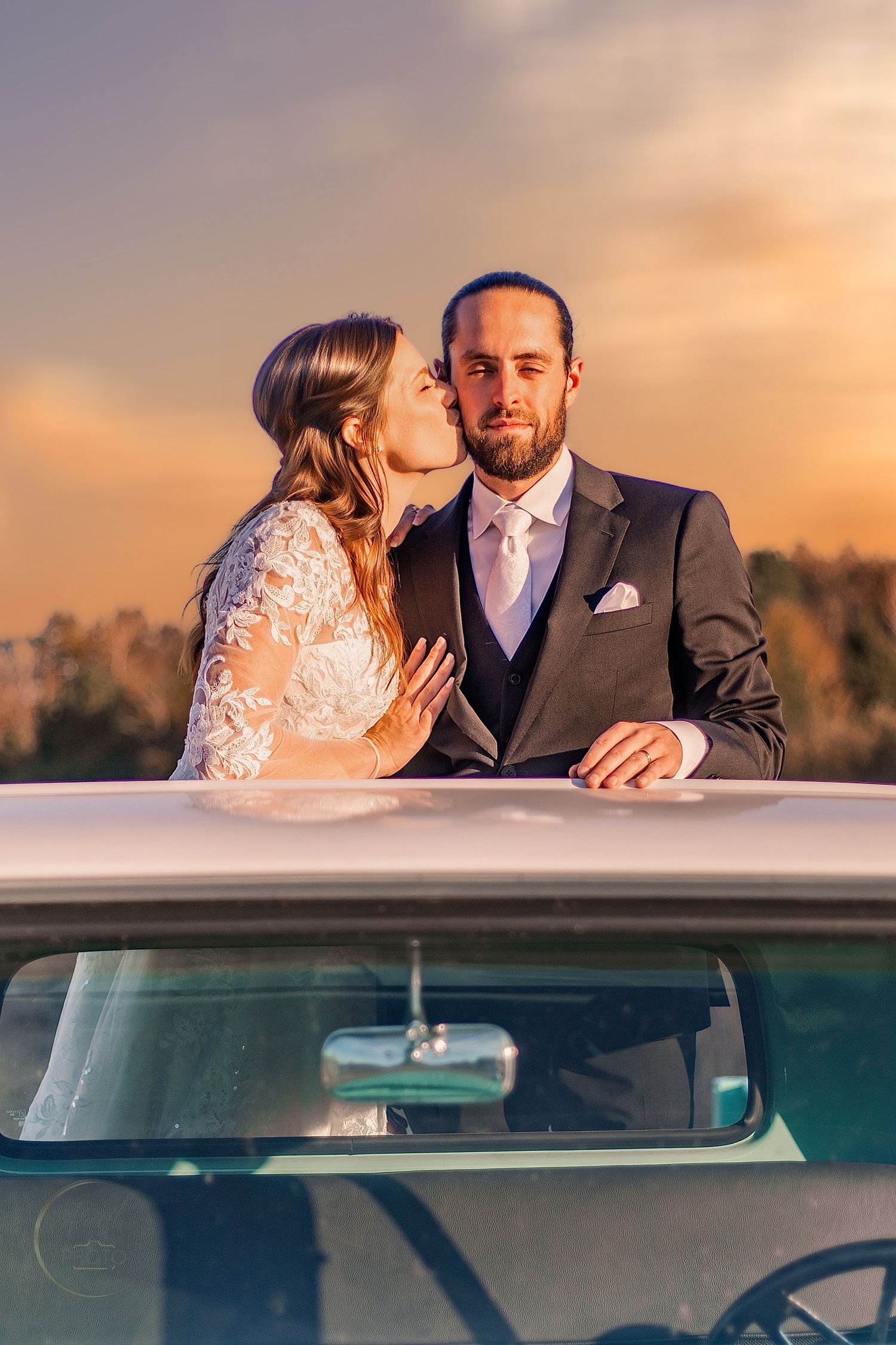 Bride kissing groom on cheek beside vintage white car at golden sunset, wedding couple in elegant attire outdoors.