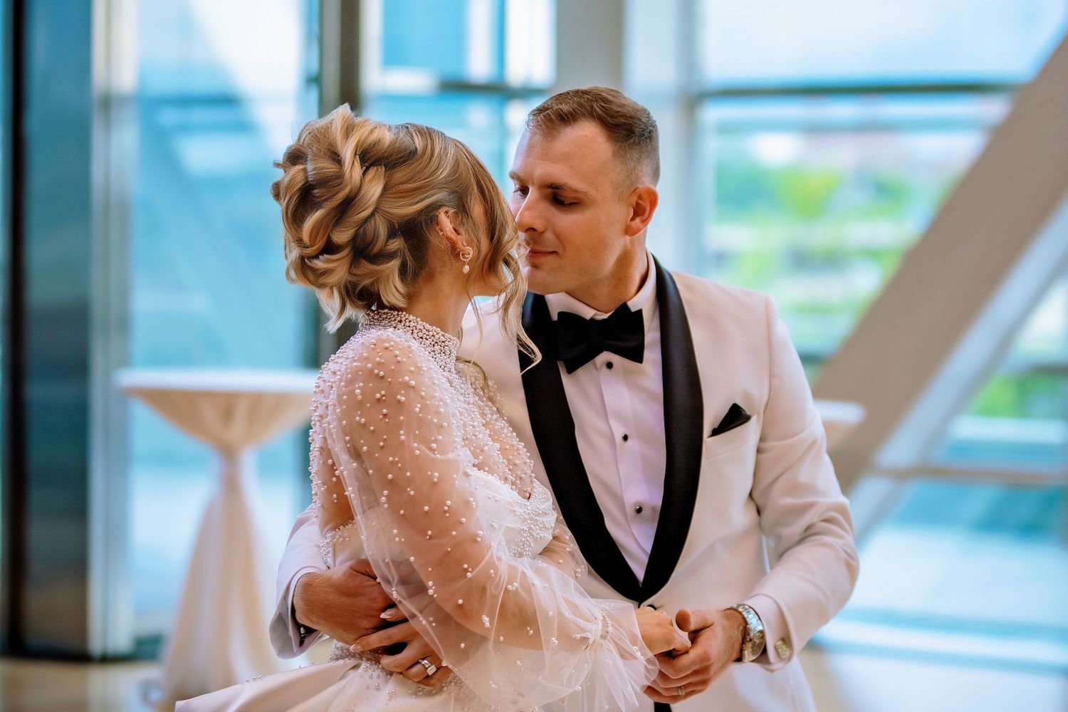 Bride and groom share an intimate first dance in a modern venue, groom in white tuxedo, bride in elegant lace gown.