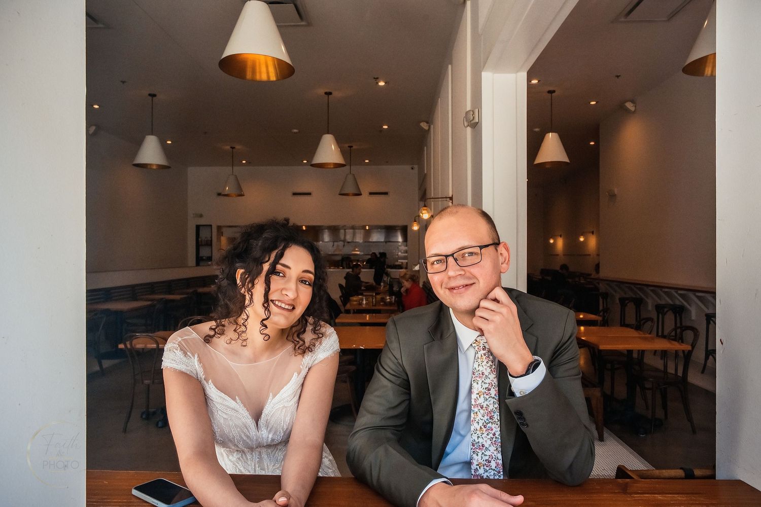 A smiling bride and groom pose together at a wooden table inside an elegant restaurant on their wedding day.