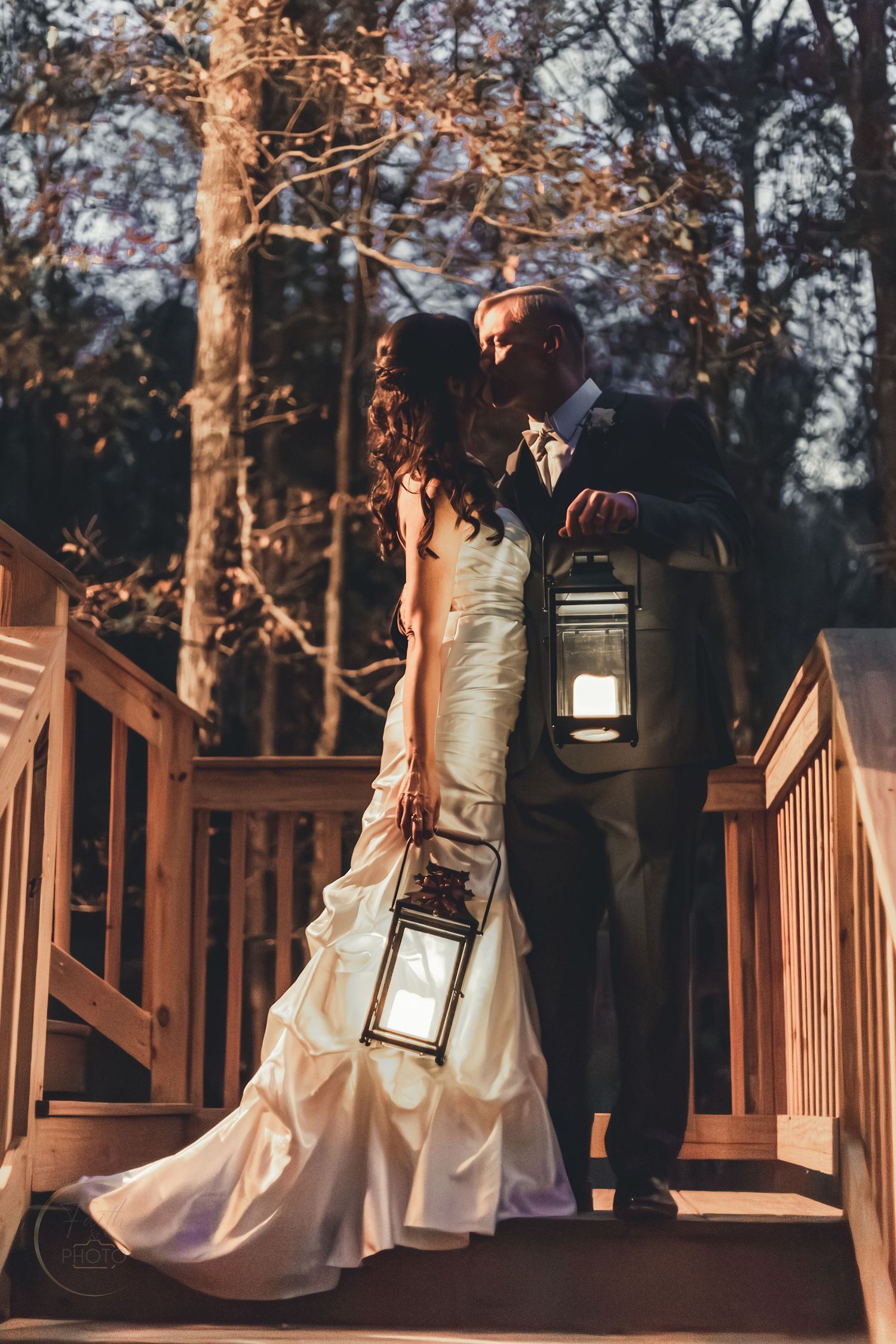 Bride and groom kissing on wooden deck steps at dusk, holding glowing lanterns, surrounded by autumn trees.