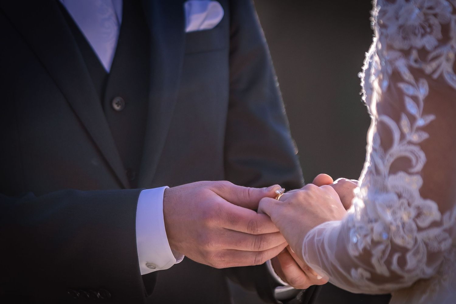 Close-up of hands exchanging wedding rings during a marriage ceremony with lace dress detail visible.