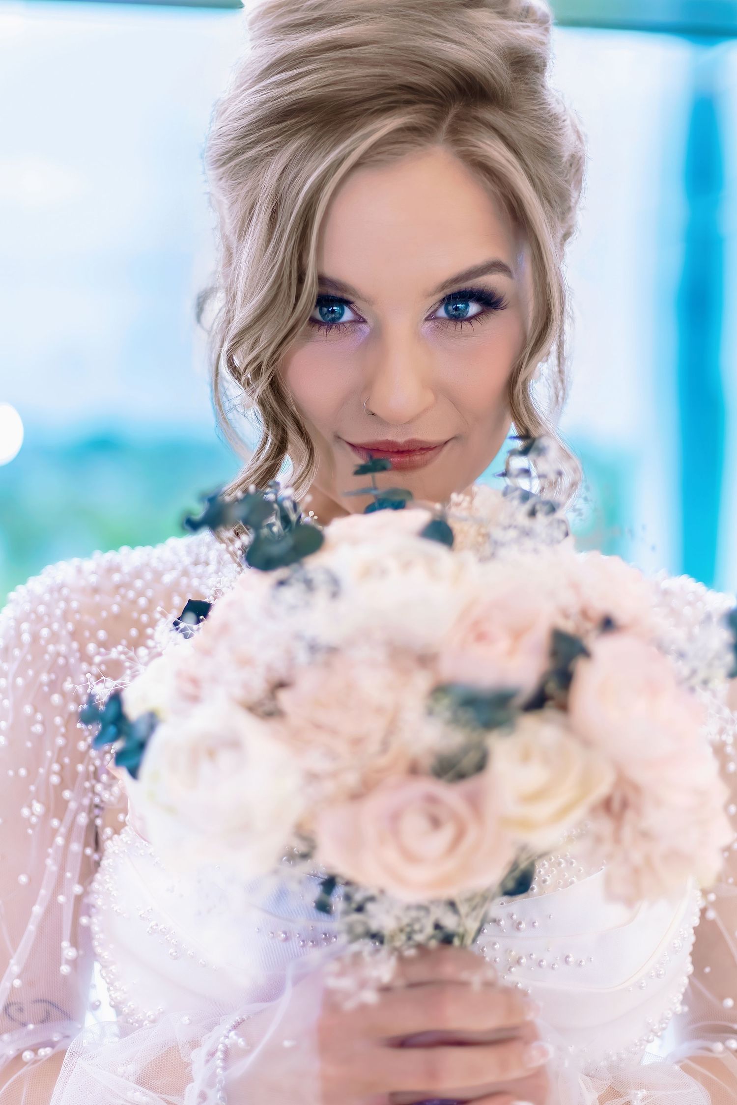 Elegant bride with blue eyes holding a bouquet of blush pink and white roses in a pearl-adorned wedding gown.