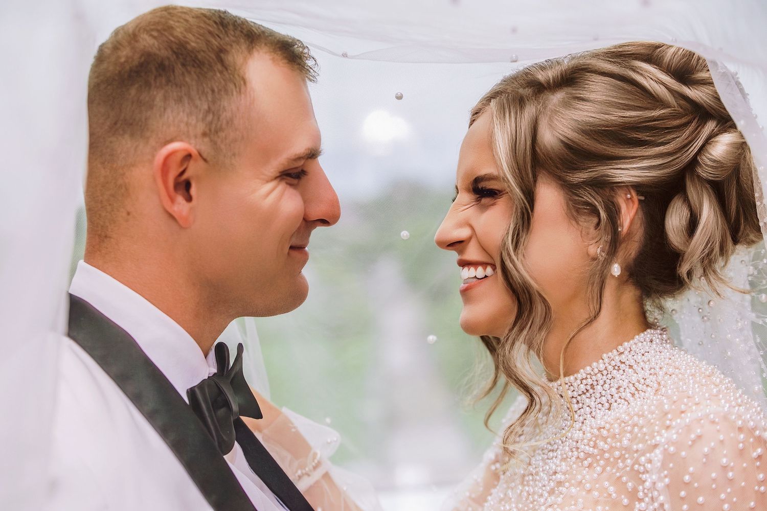 A bride and groom share an intimate moment under a veil, smiling at each other on their wedding day.
