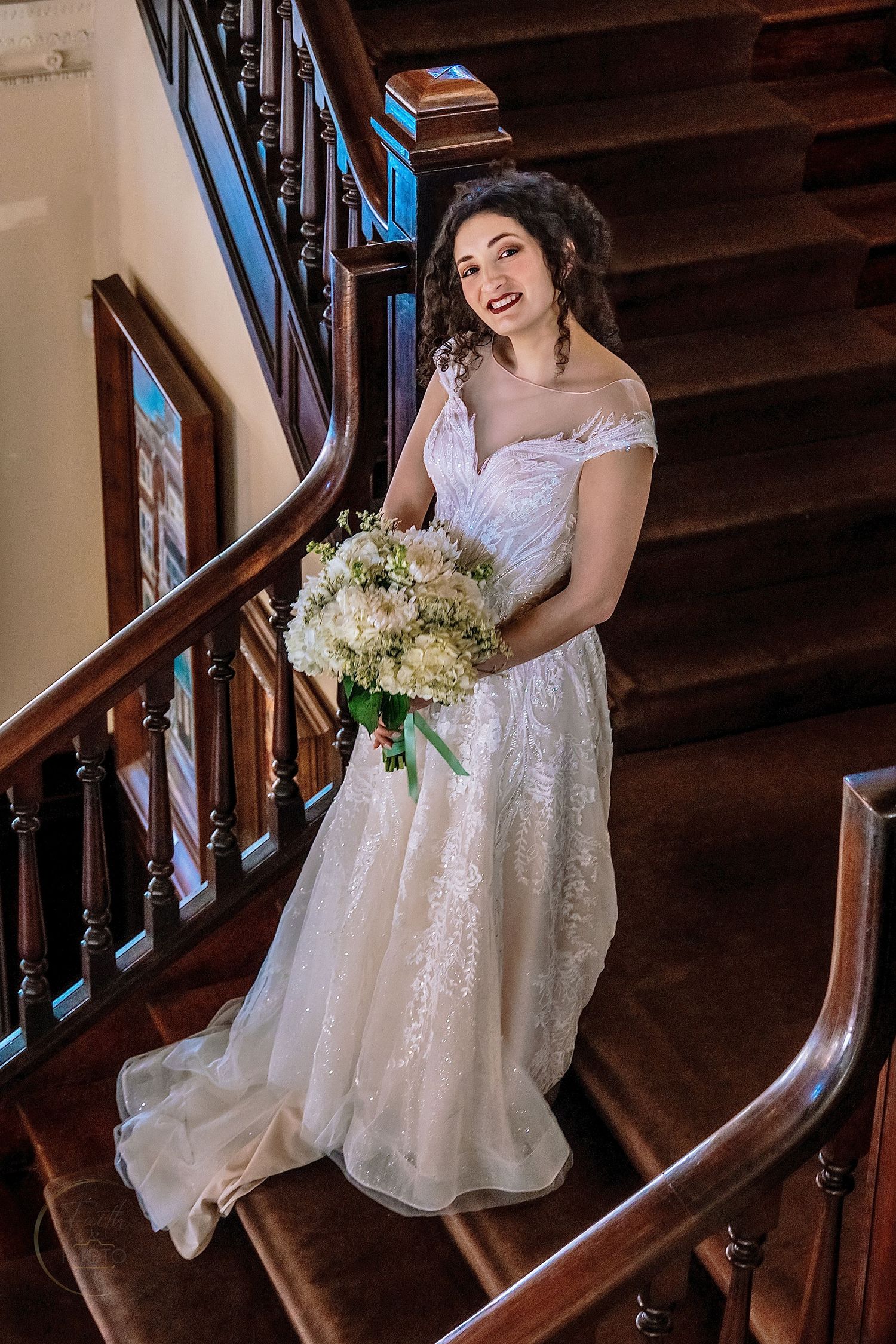 Bride in white lace off-shoulder gown holds bouquet on elegant wooden staircase, smiling radiantly.