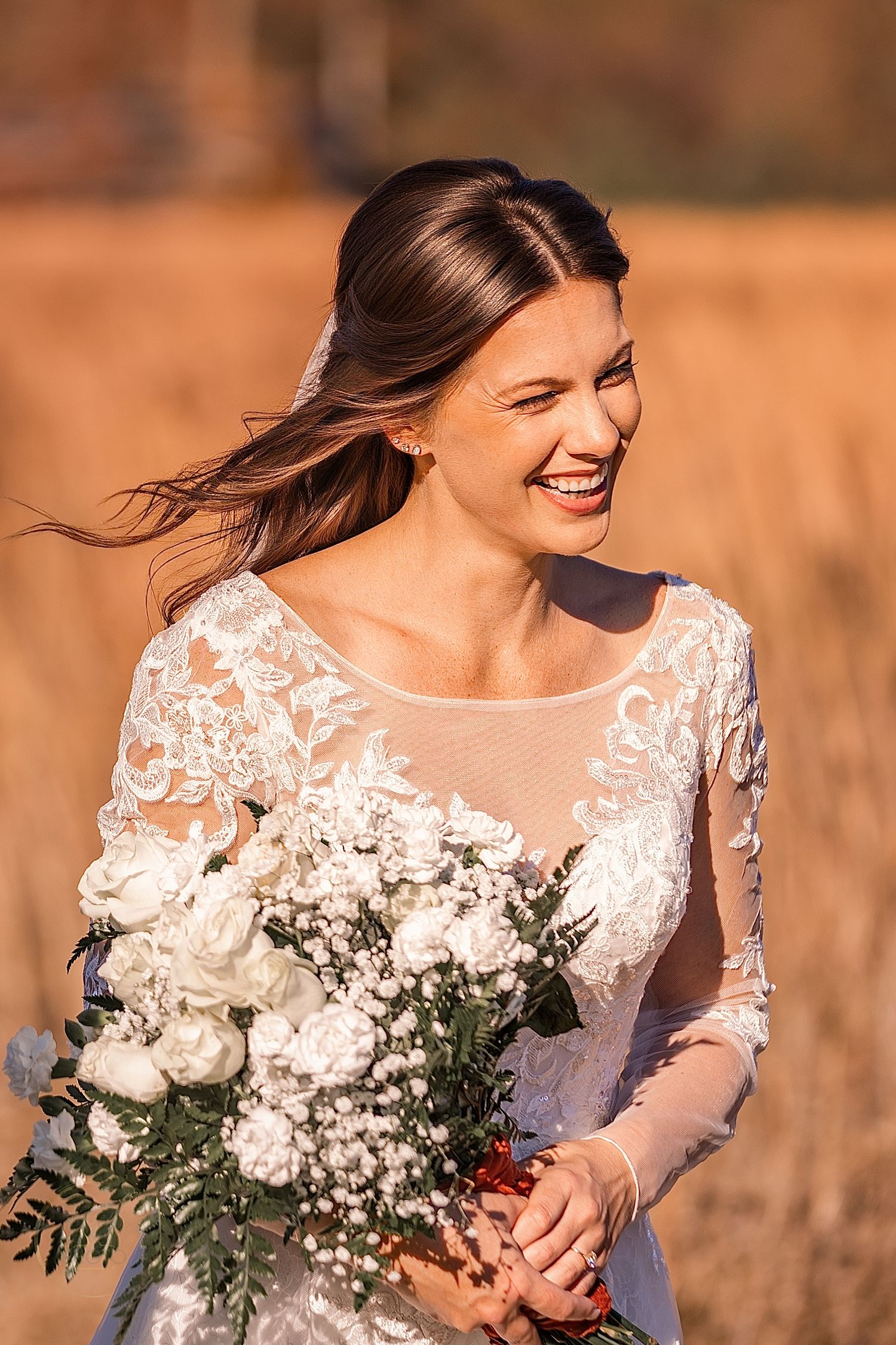 Smiling bride in lace wedding dress holding white floral bouquet outdoors in golden sunlight.
