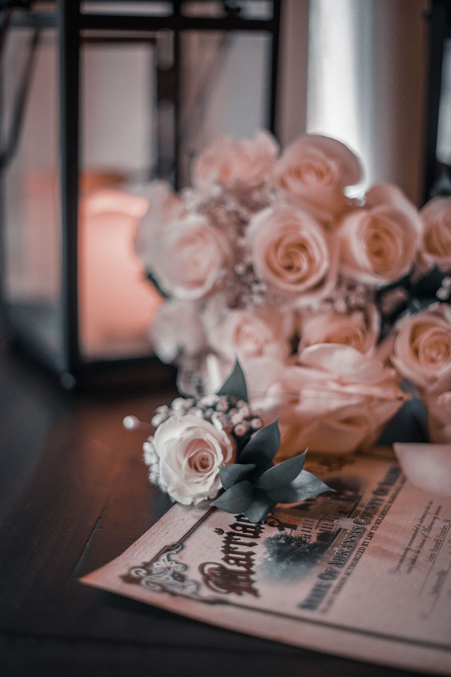 Delicate pink roses rest atop wedding invitations on a dark wooden surface in a romantic setting.