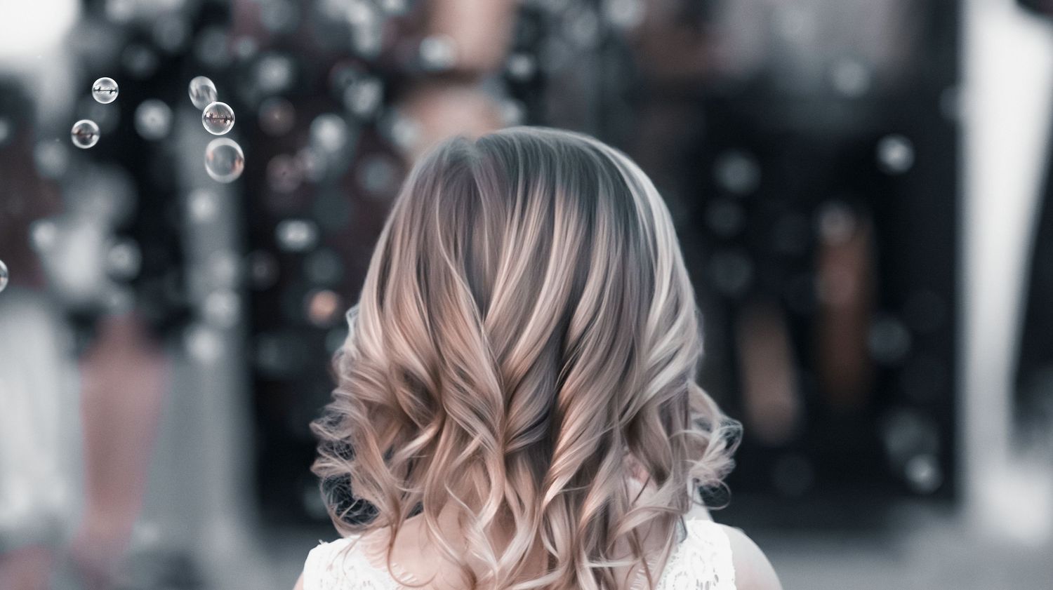 Woman with stylish ash blonde balayage wavy hair viewed from behind, with bokeh bubbles in the background.
