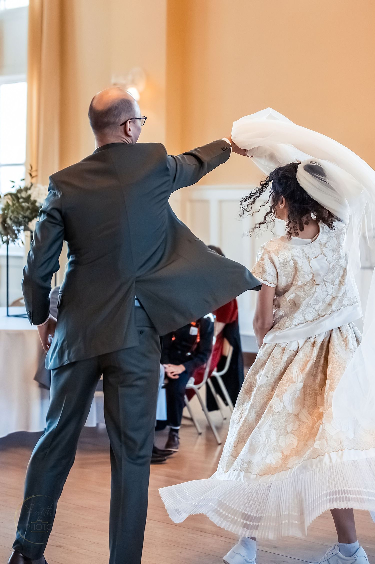 Father spinning bride in elegant gold dress and veil during wedding reception dance in a warmly lit ballroom.