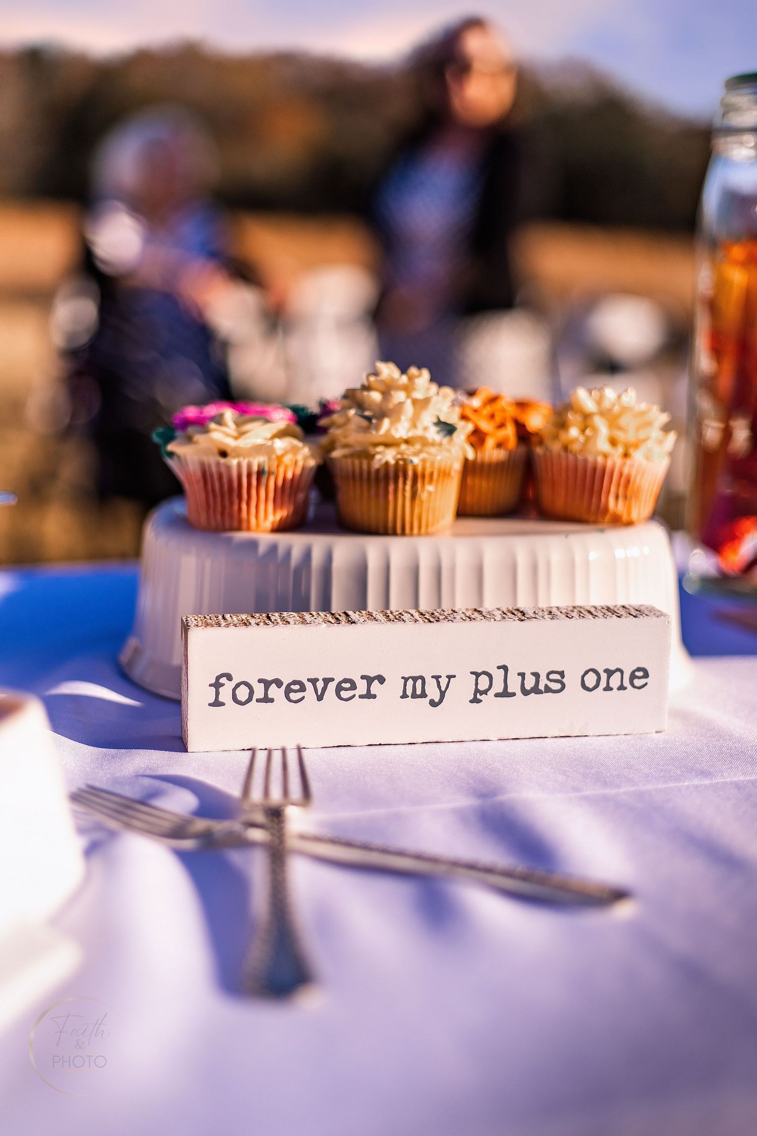Wedding cupcake stand with forever my plus one sign on white tablecloth at outdoor reception.