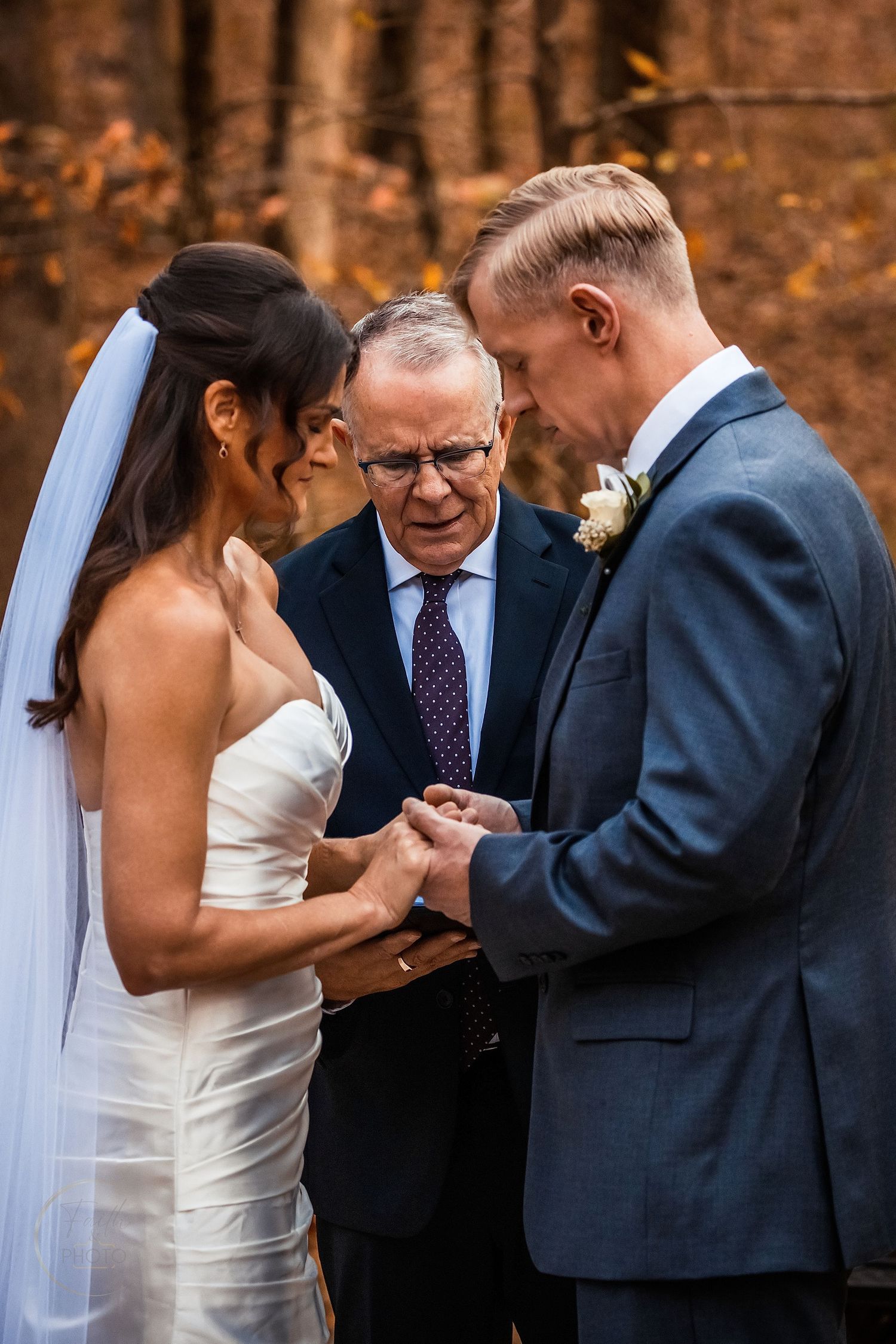 Bride and groom exchange rings during outdoor fall wedding ceremony with officiant in autumn forest setting.