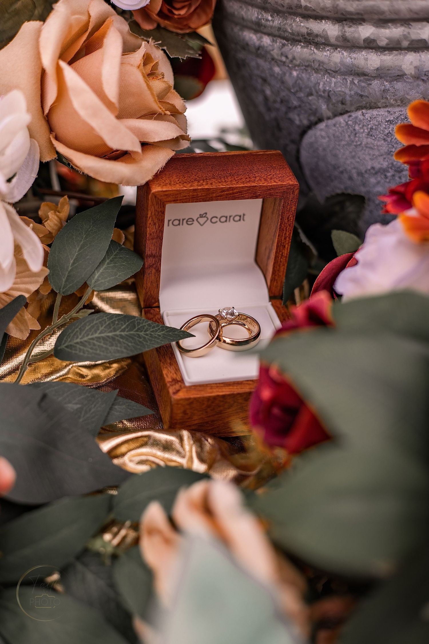 Gold wedding rings displayed in a Rare Carat wooden box surrounded by elegant floral arrangements.