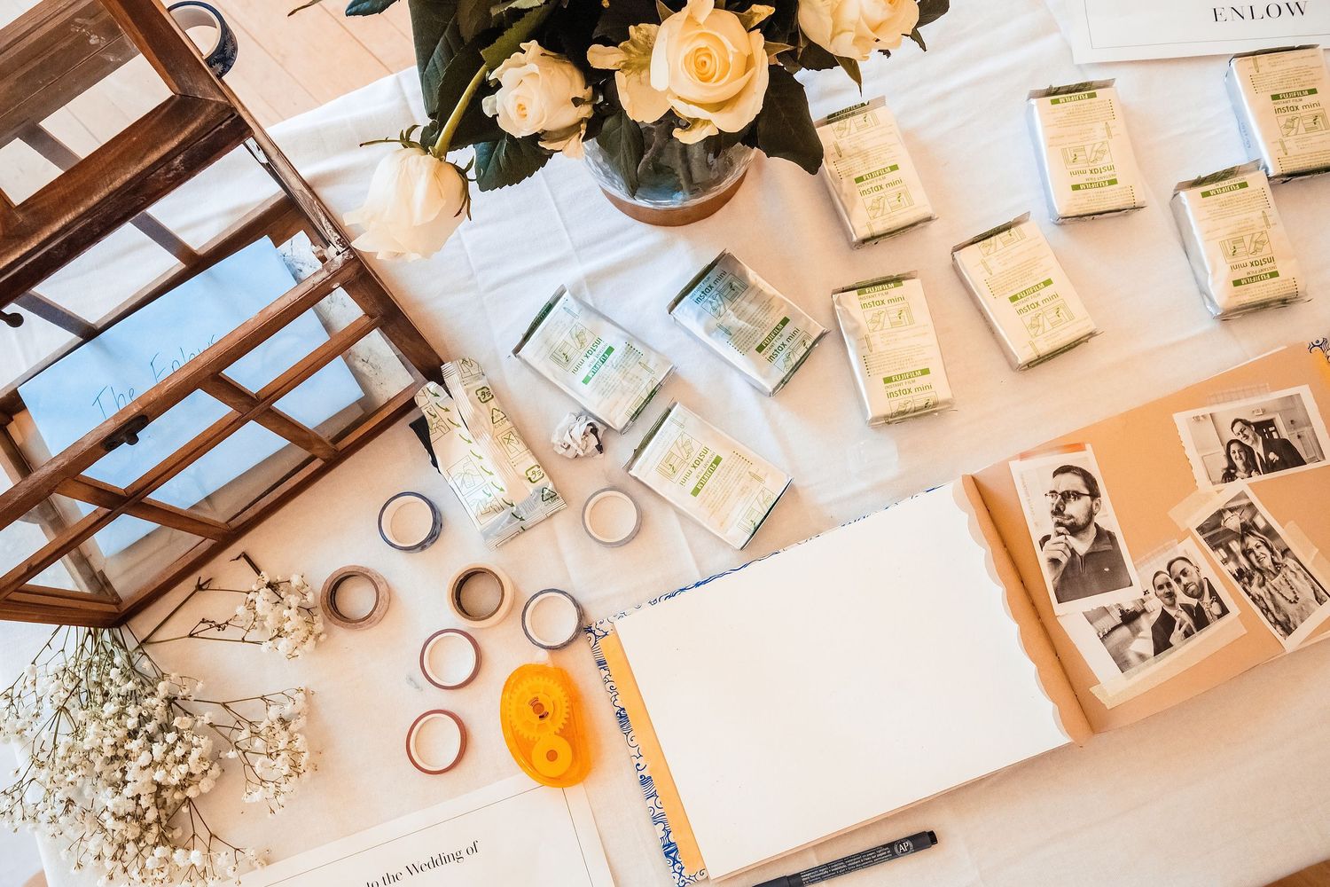 Overhead view of a wedding craft table with washi tape, rings, an open guest book, photos, and floral arrangements.