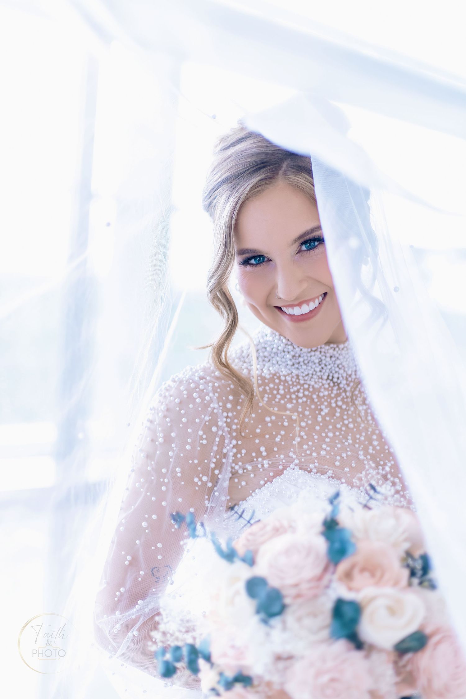 Smiling bride in pearl-embellished wedding gown holds blush bouquet under flowing veil in bright natural light.