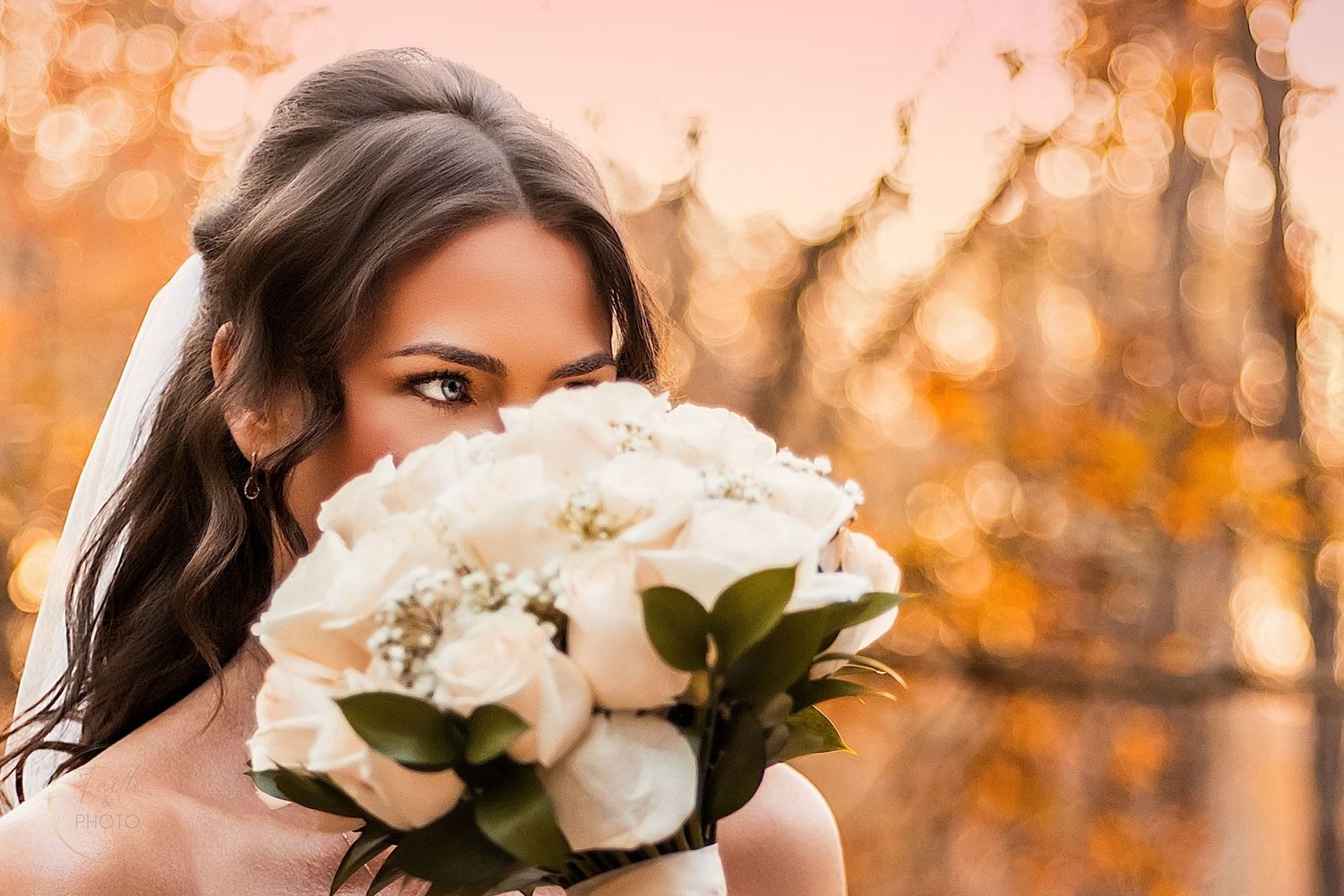 Bride in white veil holding bouquet of white roses, with autumn foliage bokeh background.
