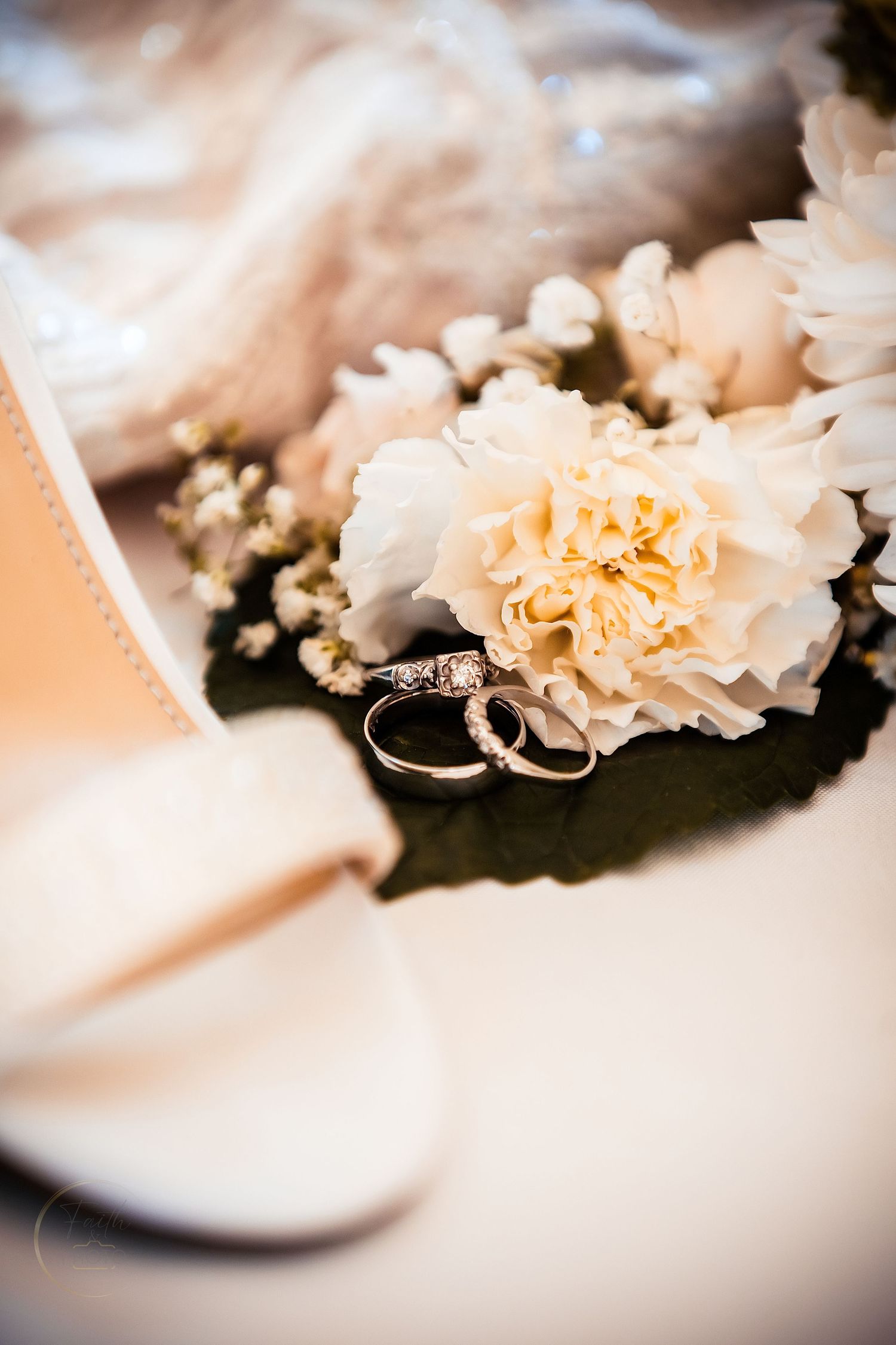 Two gold wedding rings rest on white carnation flowers beside bridal shoes and veil.