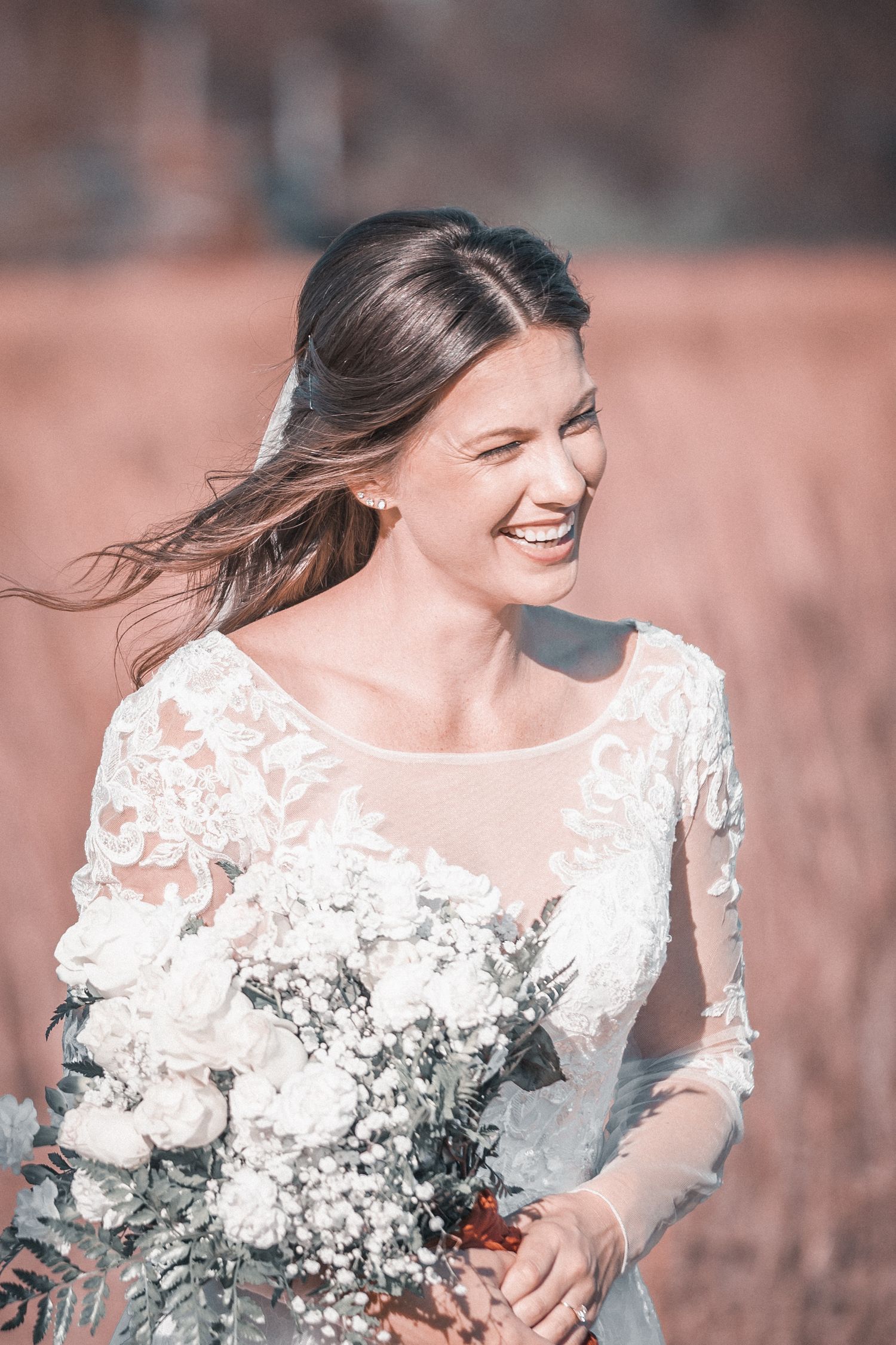 A bride in a delicate white lace wedding dress smiles joyfully while posing outdoors in natural lighting.