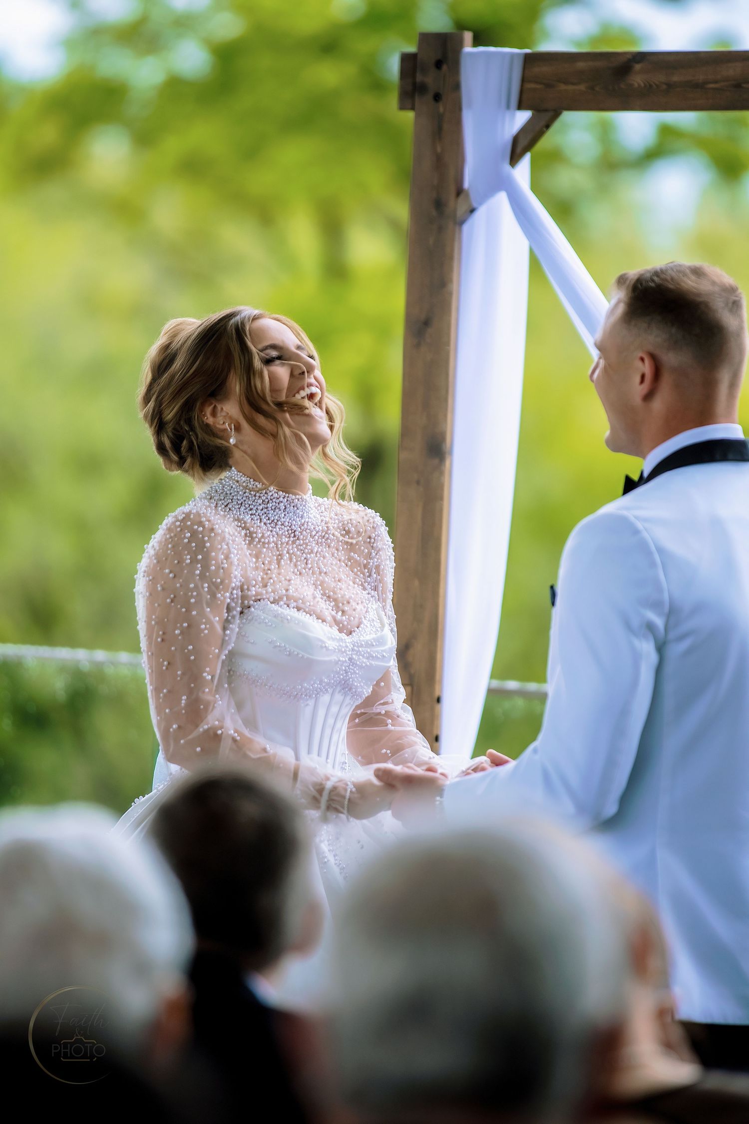 Bride in pearl-embellished white gown laughing joyfully with groom during outdoor wedding ceremony under wooden arch.