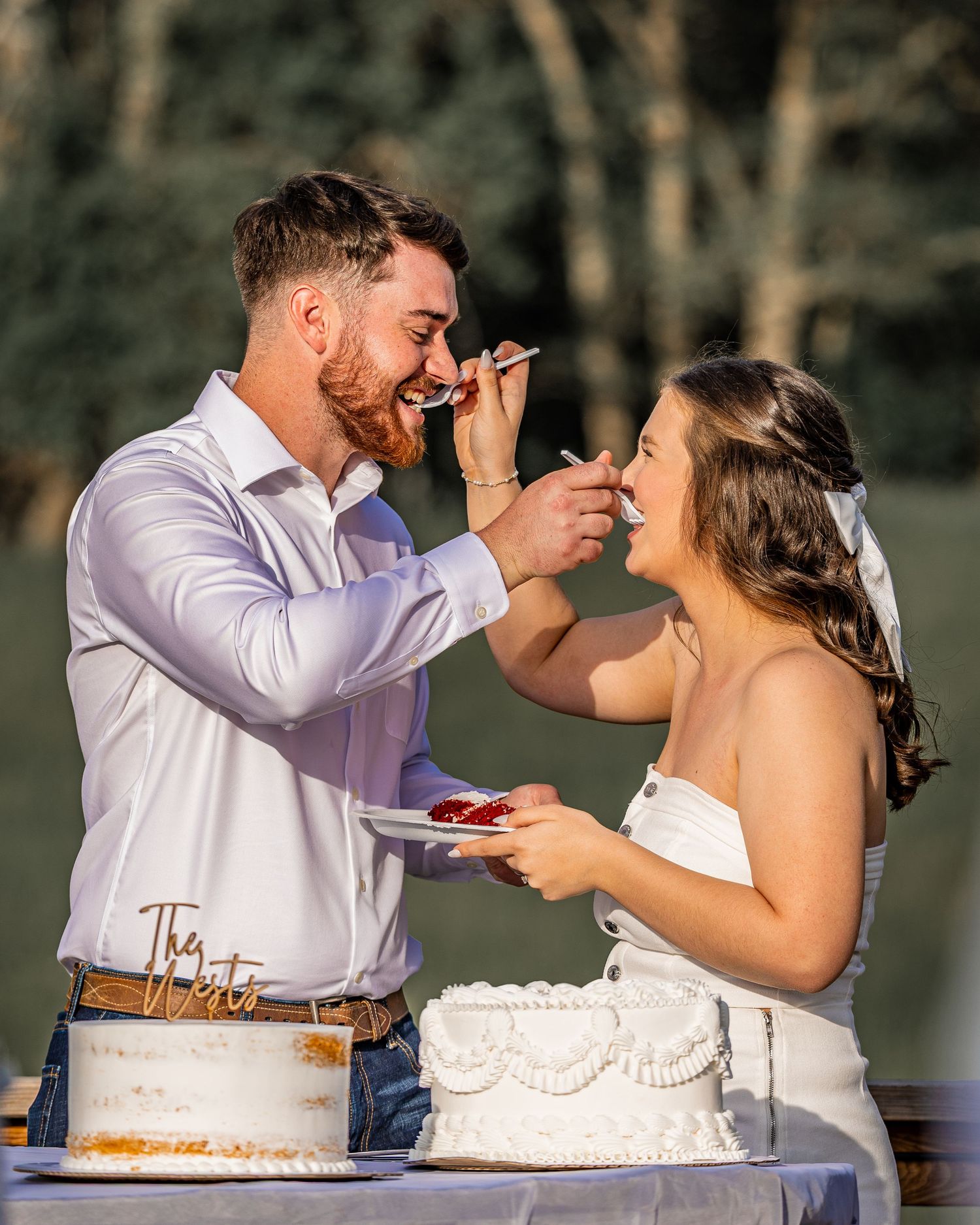 A newlywed couple playfully smears wedding cake on each other's faces during their outdoor reception celebration.