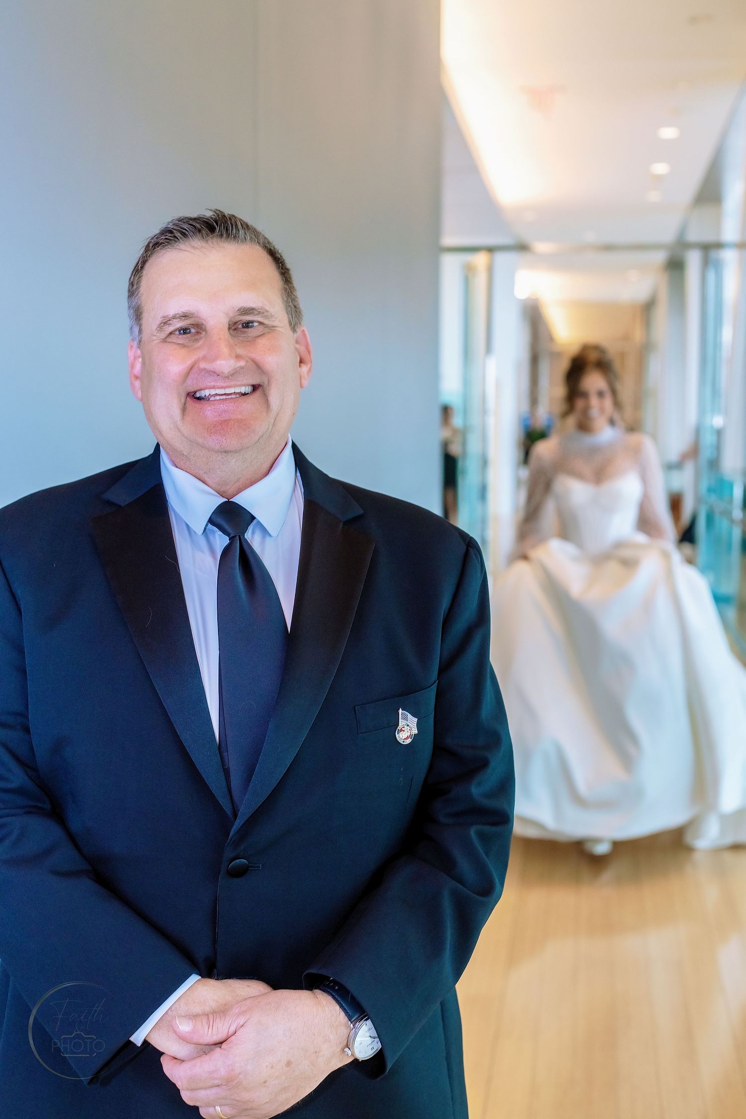 Man in tuxedo smiles as bride in white gown approaches down hallway at elegant wedding venue.