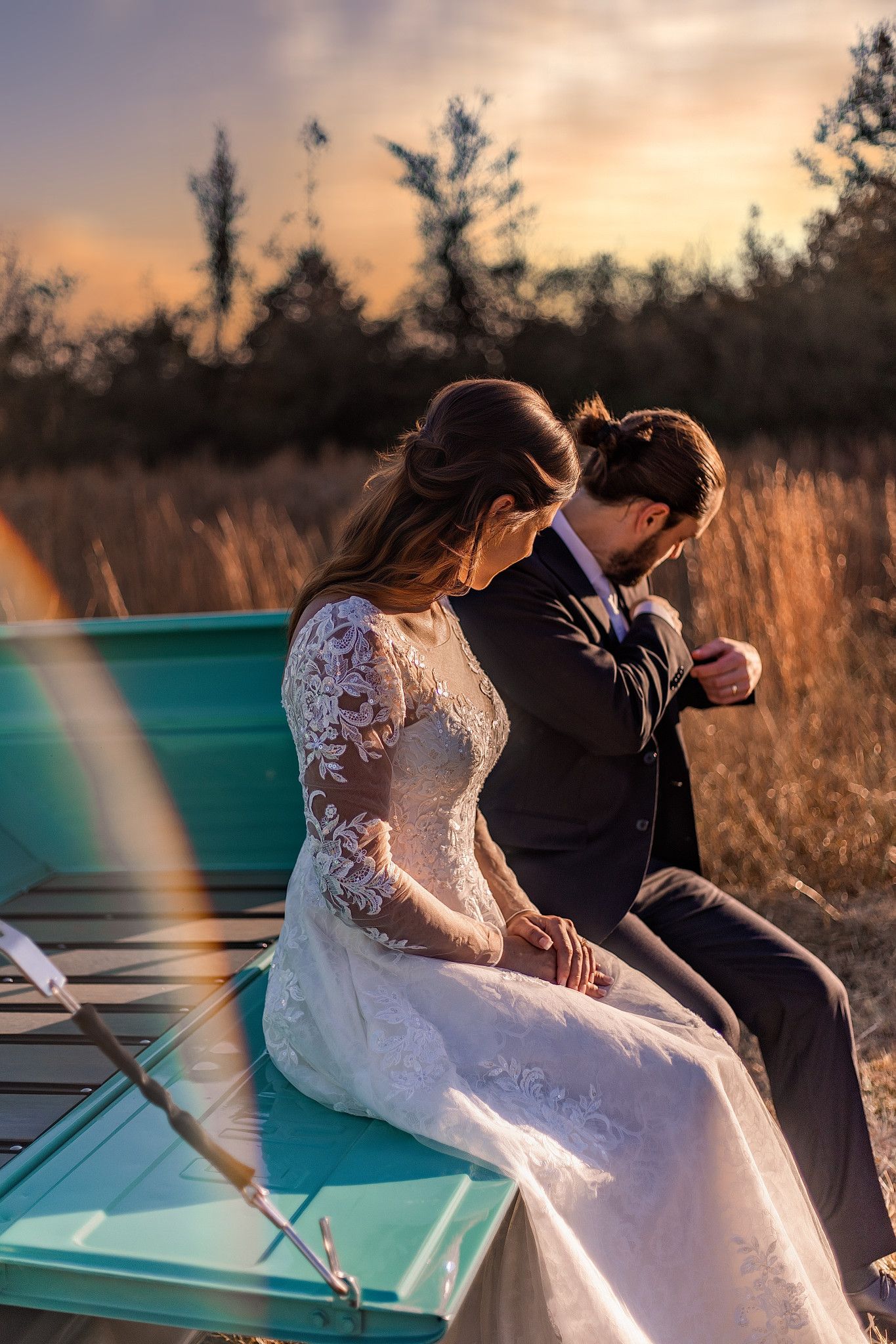 Bride and groom share intimate moment sitting on turquoise boat at golden sunset, surrounded by autumn fields.