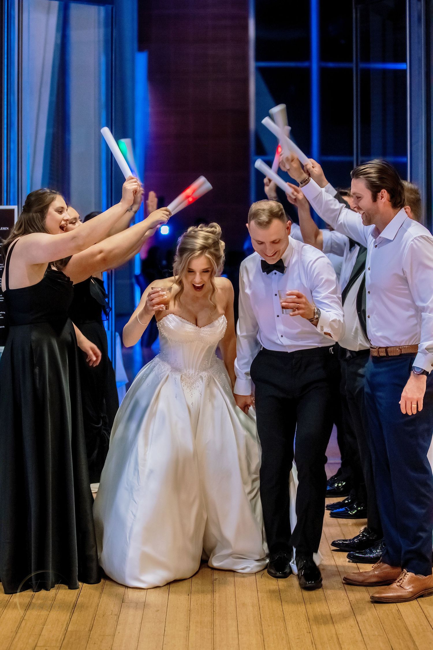 Bride and groom walk through a glowstick tunnel surrounded by cheering guests at an elegant wedding reception.