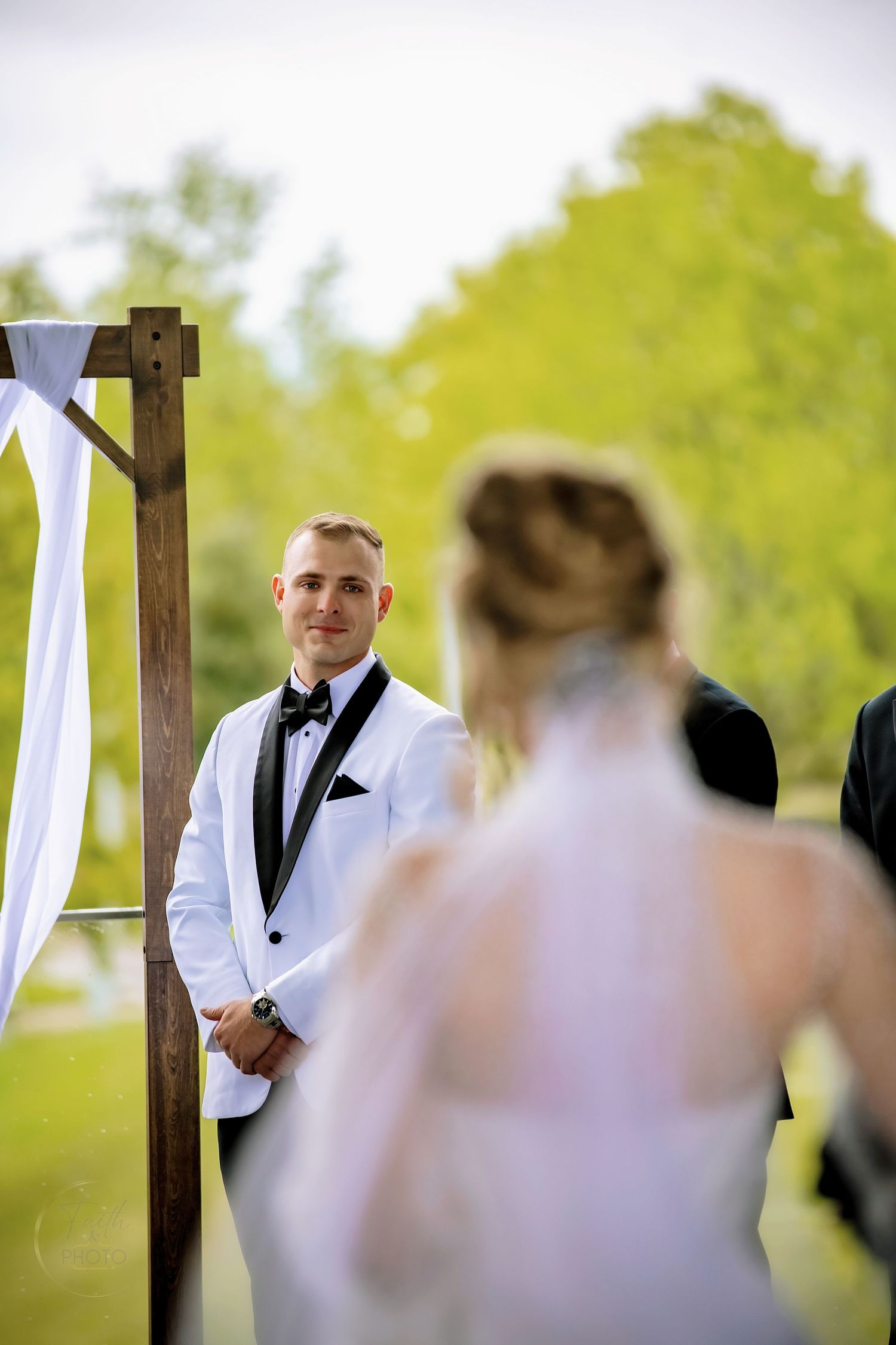 Groom in white tuxedo smiles at bride approaching outdoor wedding altar with wooden arch and white draping.